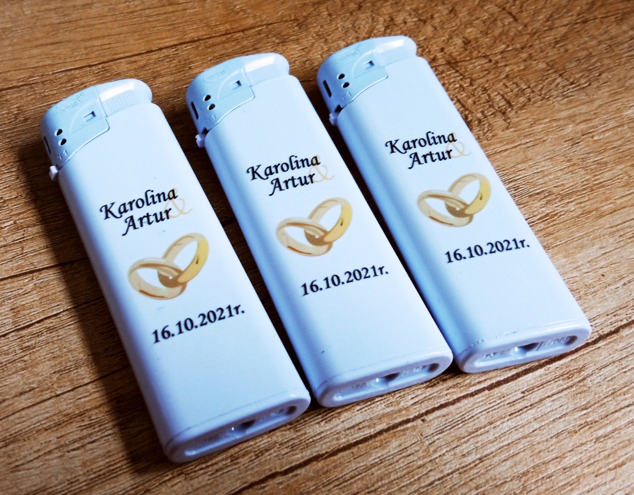 Trzy zapalniczki z nadrukiem imion Karolina i Artur, data 16.10.2021 oraz grafika złotych obrączek na drewnianym tle. Pamiątka ślubna.