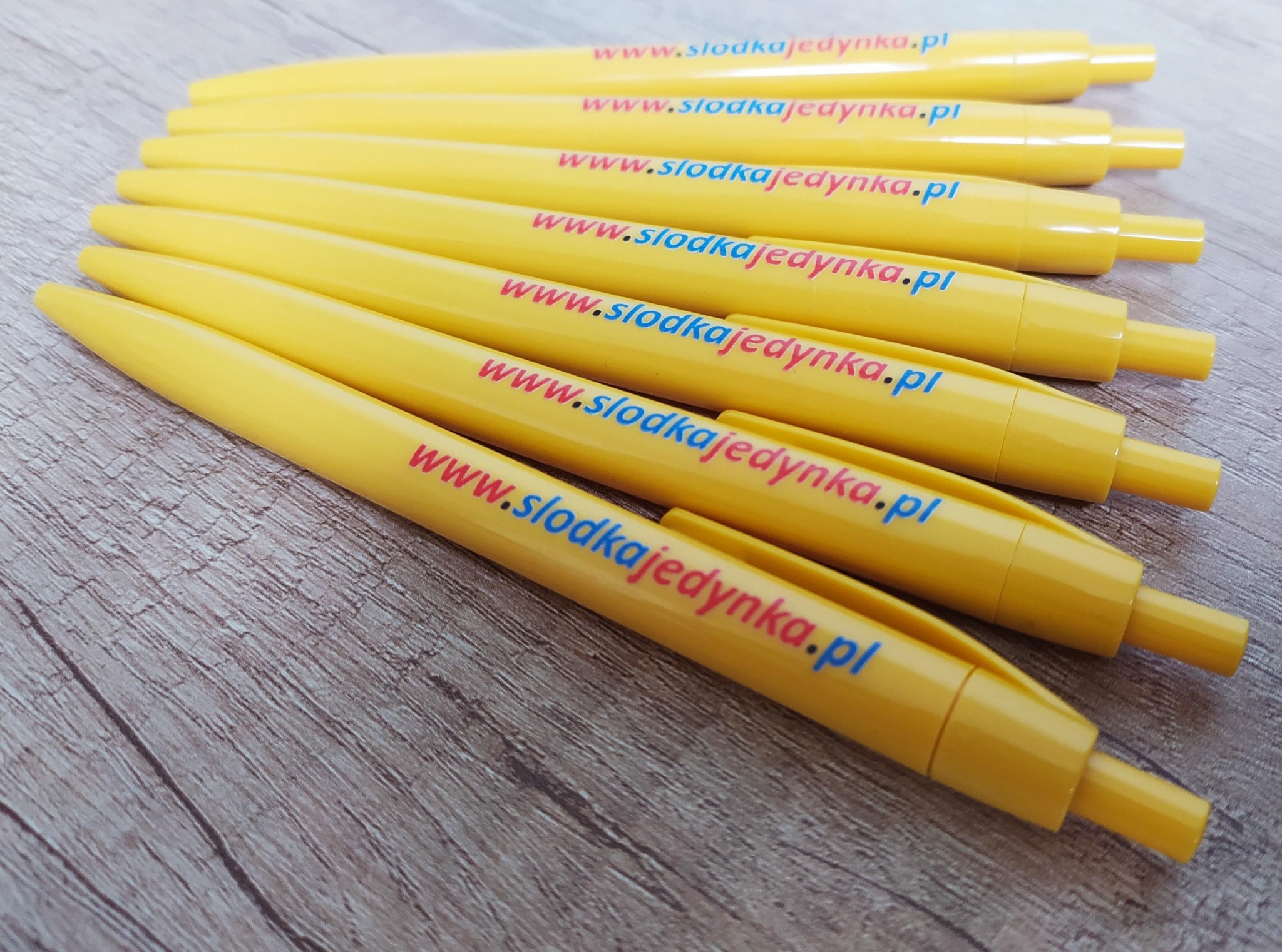 Żółte długopisy z nadrukowanym kolorowym logo 'www.slodkajedynka.pl' ułożone równolegle na drewnianym tle. Widoczne detale nadruku i faktura długopisów.