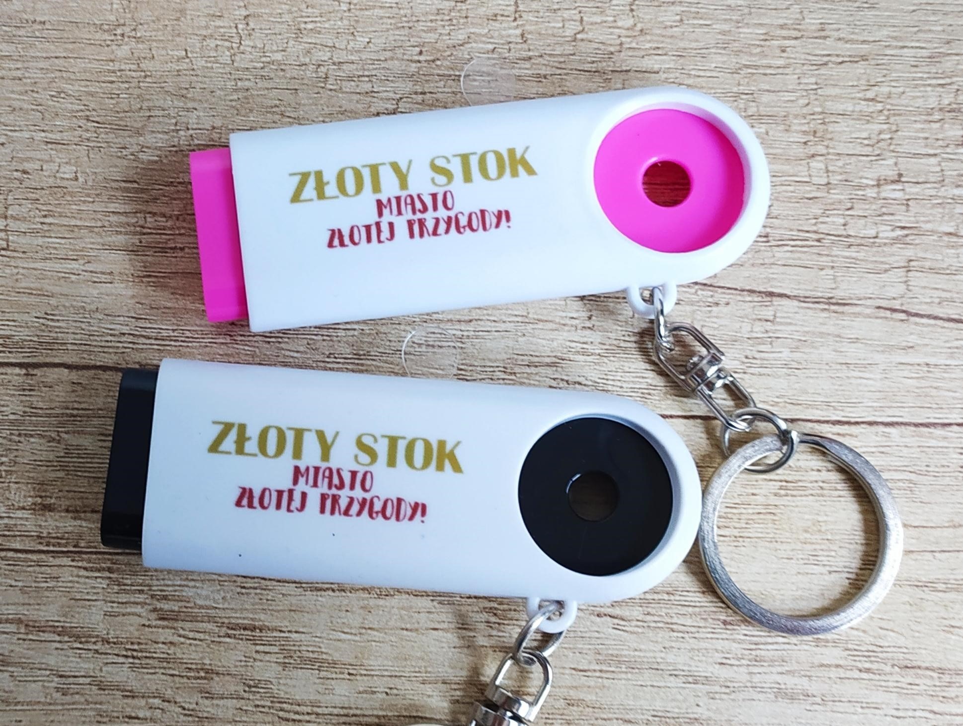 Dwa białe breloczki z napisem 'Złoty Stok miasto złotej przygody!' na drewnianym tle. Jeden z różowym, drugi z czarnym elementem.