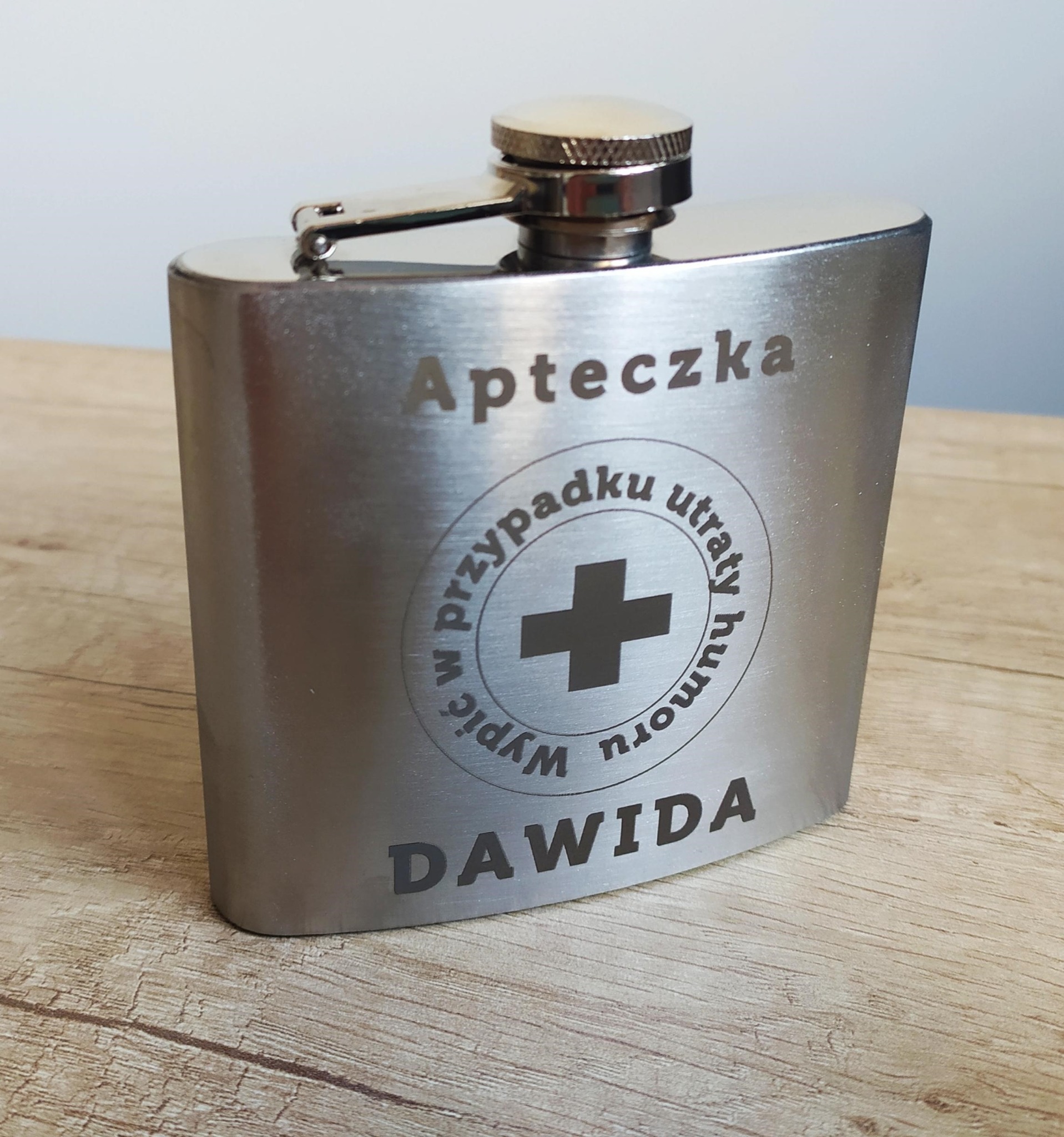 Metalowa piersiówka z grawerem 'Apteczka w przypadku utraty humoru' i imieniem 'DAWIDA', leżąca na drewnianym blacie. Prezent z personalizacją.