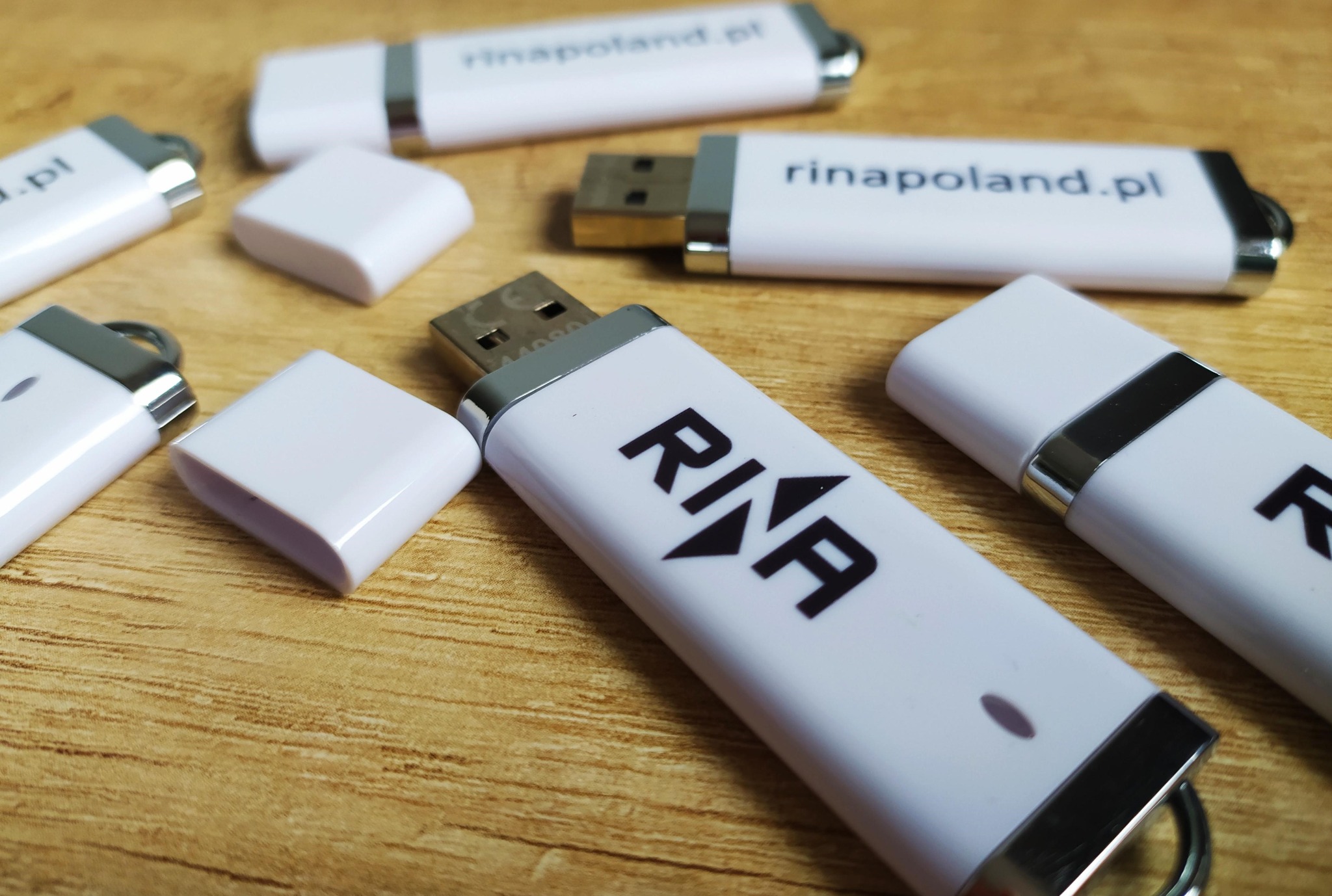 Białe pendrive z logo firmy RINA i adresem rinapoland.pl leżące na drewnianym stole. Widoczne złącza USB i nakładki. Prezentacja identyfikacji wizualnej.