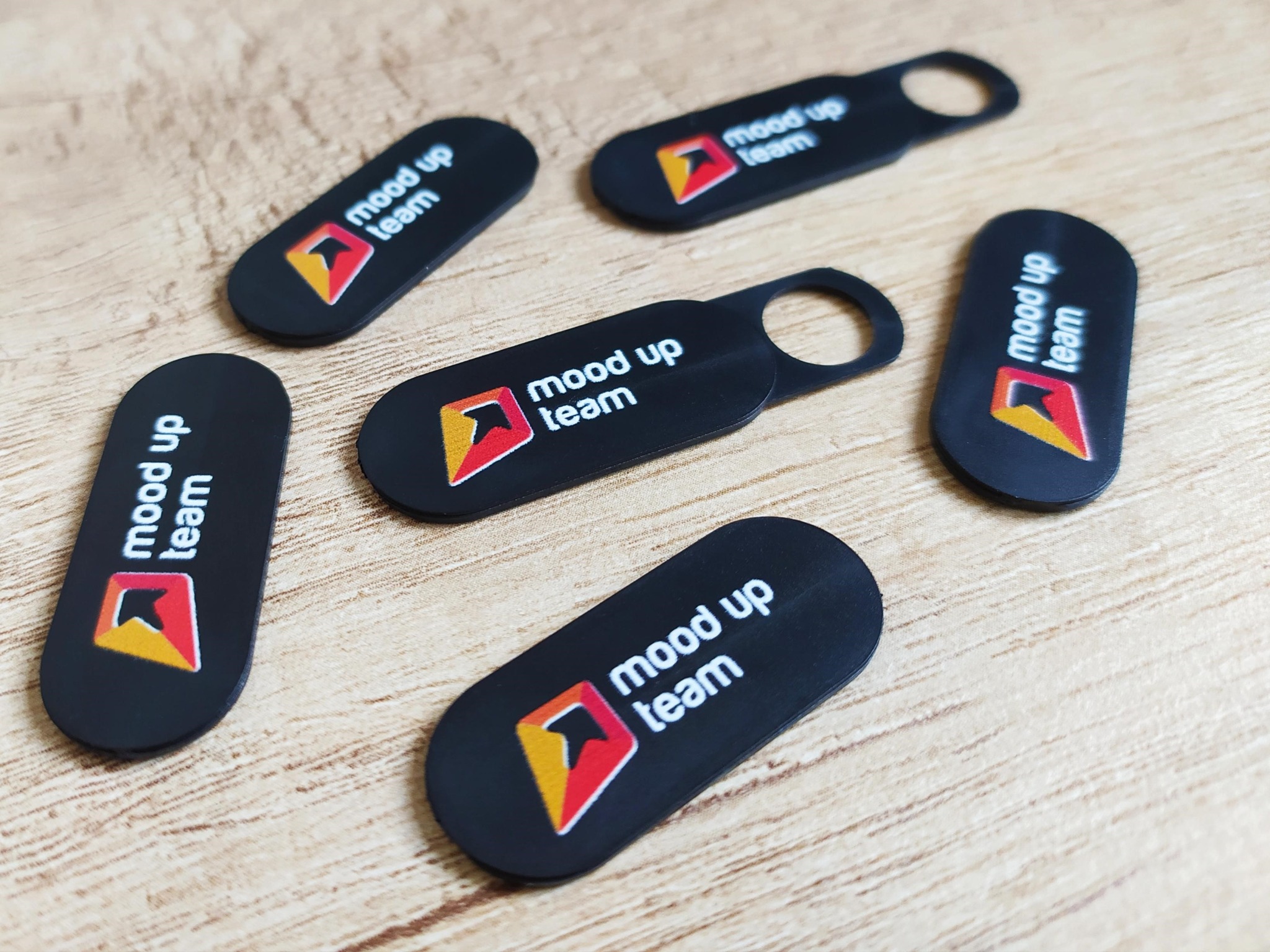 Sześć czarnych, podłużnych breloków z logo 'mood up team' leżących na drewnianym stole. Breloki mają zaokrąglone rogi i otwór na kółko.
