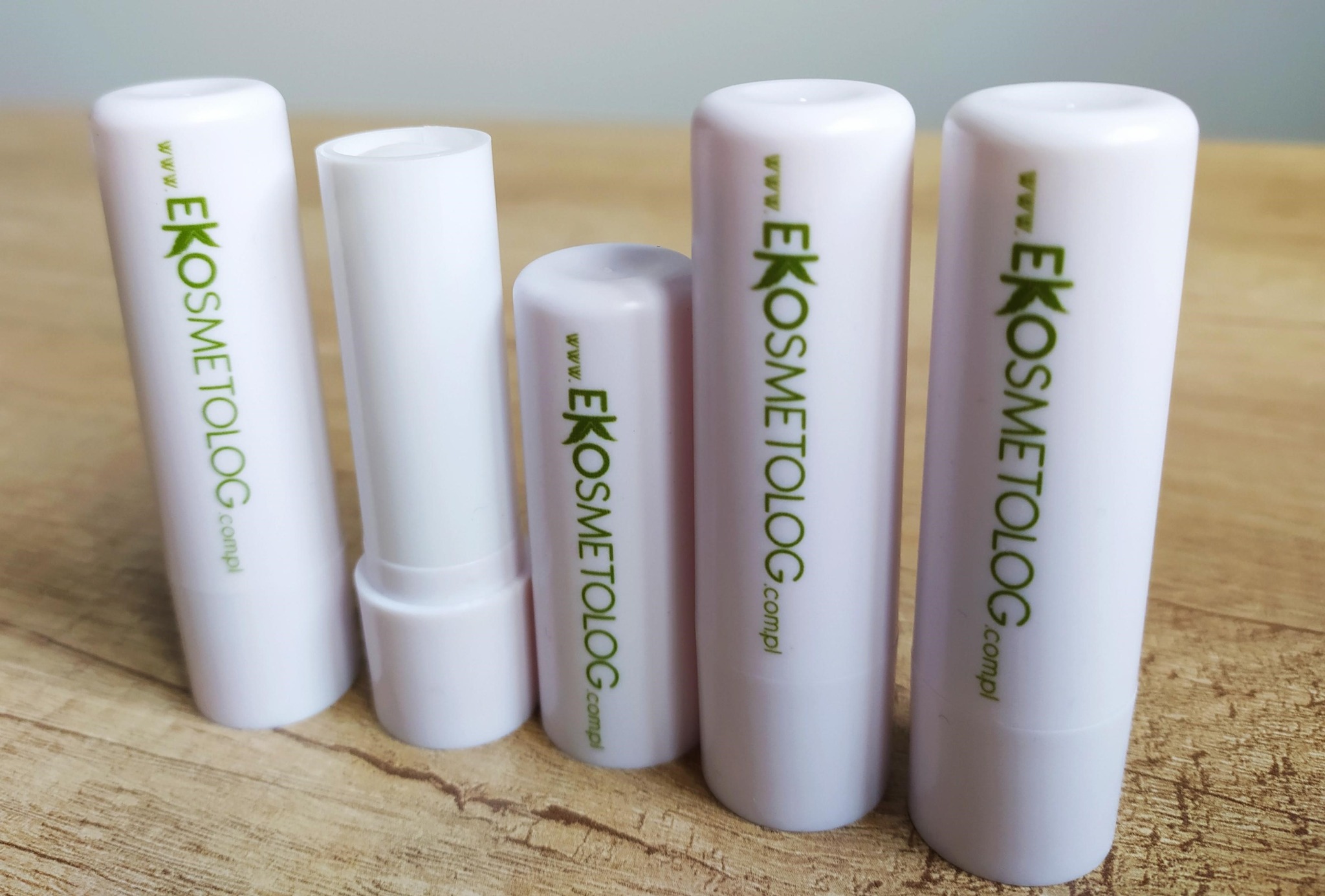 Pięć białych pomadek ochronnych Eko Kosmetolog o różnej wysokości, ustawionych na drewnianym blacie, z zielonym logo i adresem strony internetowej.