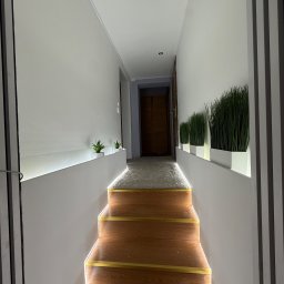 Norbert Mazur - Nowoczesny korytarz z oświetleniem LED wzdłuż schodów i ścian, minimalistyczne donice z roślinami, świeżo pomalowane ściany w odcieniach bieli.