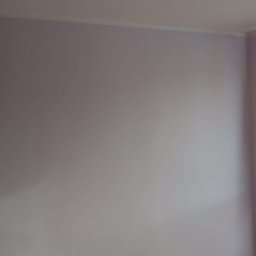 Norbert Mazur - Gładka, pomalowana na jasny odcień fioletu ściana w rogu pomieszczenia, z widoczną listwą przypodłogową przy suficie. Minimalistyczne wykończenie wnętrza.