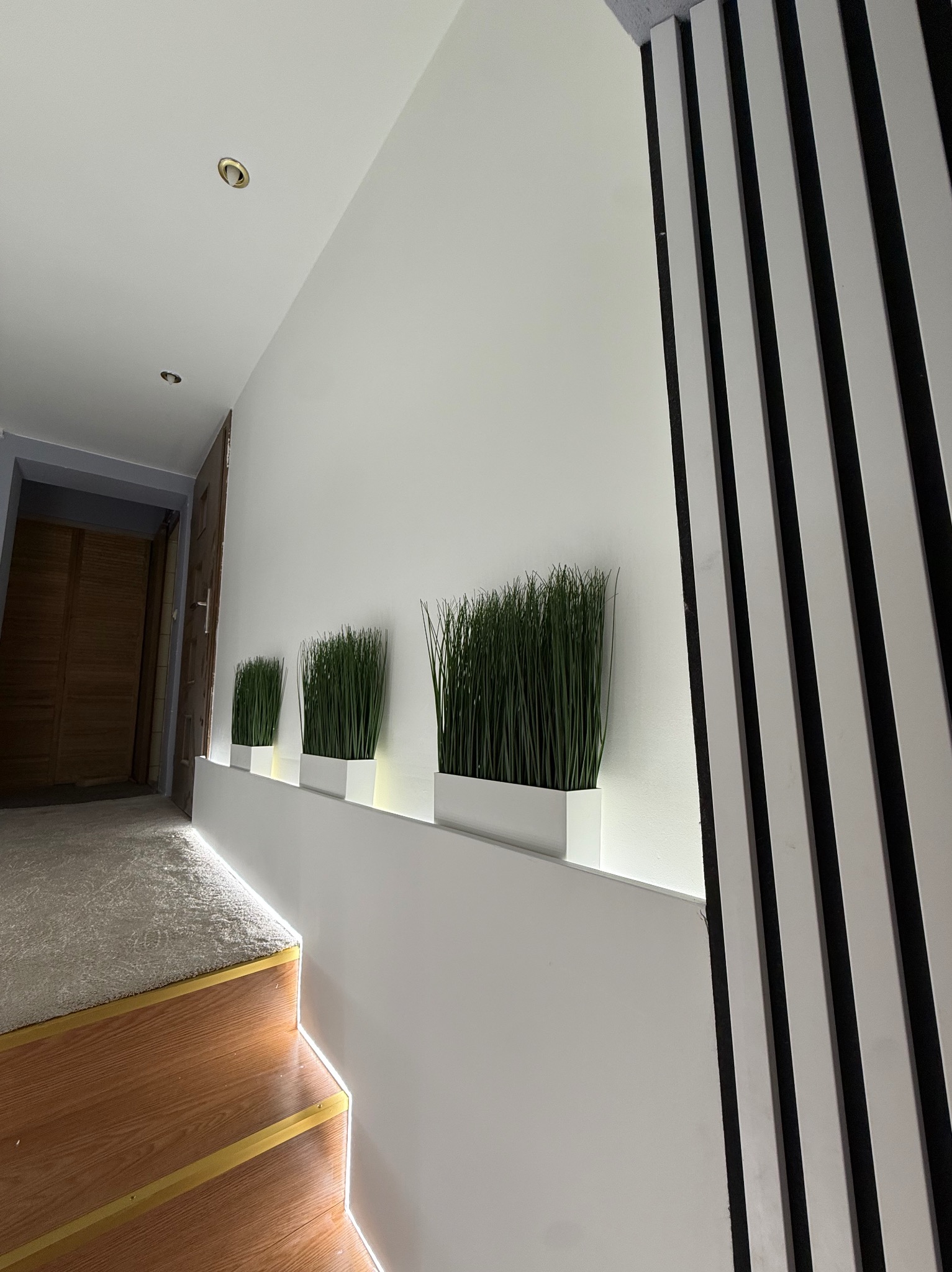 Biała ściana z trzema donicami z zieloną trawą, podświetlona listwą LED. Obok drewniane schody z oświetleniem i dekoracyjne panele ścienne w czarno-białe pasy.