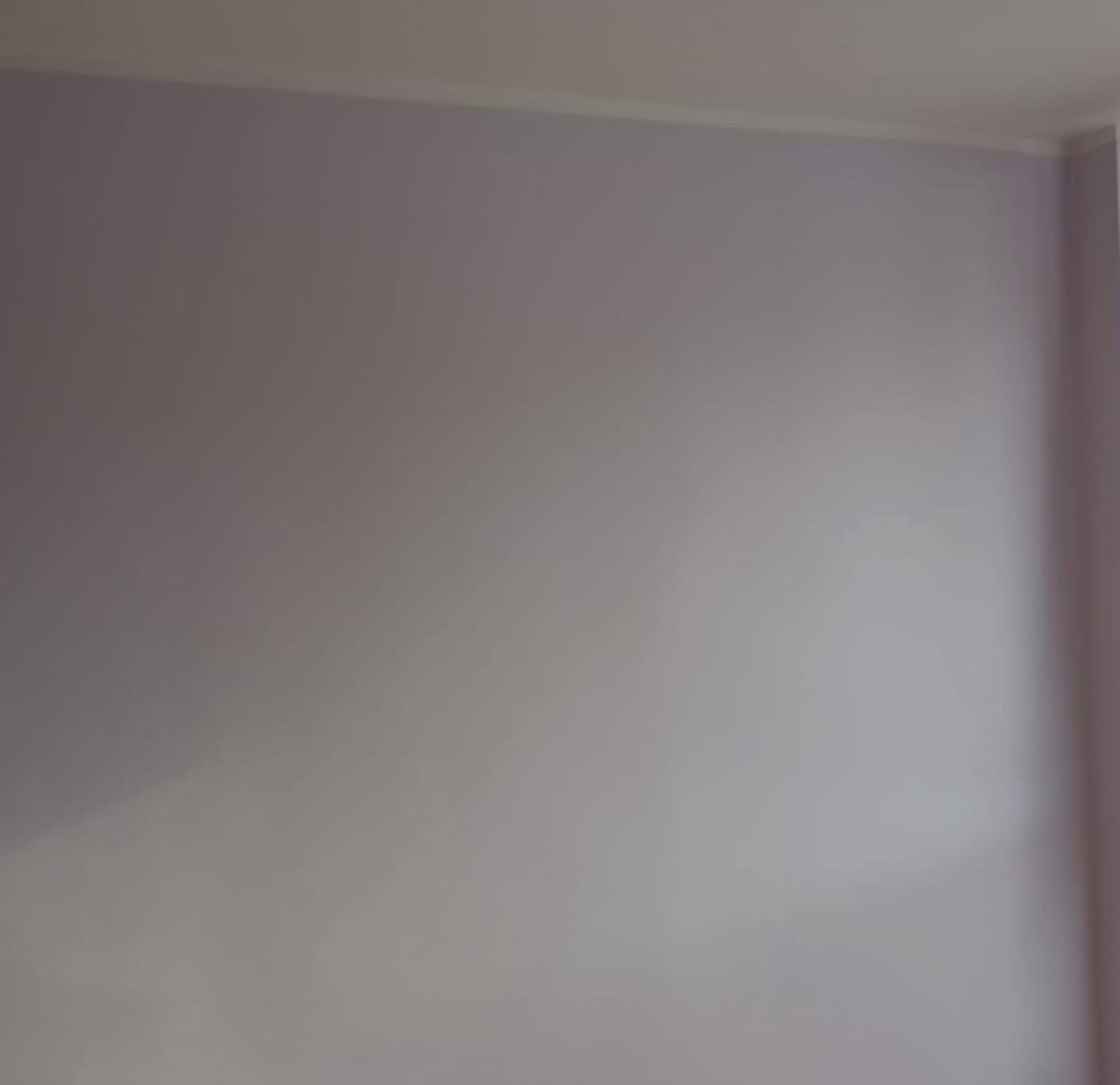 Gładka, pomalowana na jasny odcień fioletu ściana w rogu pomieszczenia, z widoczną listwą przypodłogową przy suficie. Minimalistyczne wykończenie wnętrza.