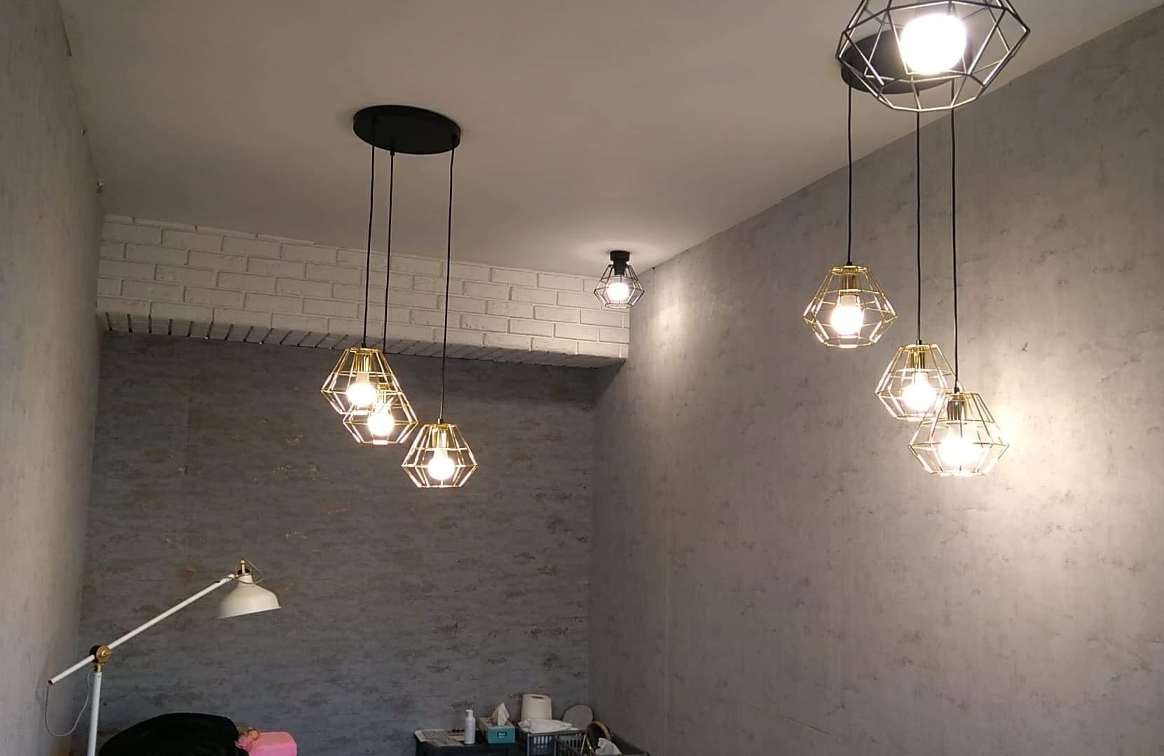 Ściana z efektem dekoracyjnym w odcieniach szarości, oświetlona przez wiszące lampy z geometrycznymi kloszami. Widoczny fragment podłogi i lampa stojąca.