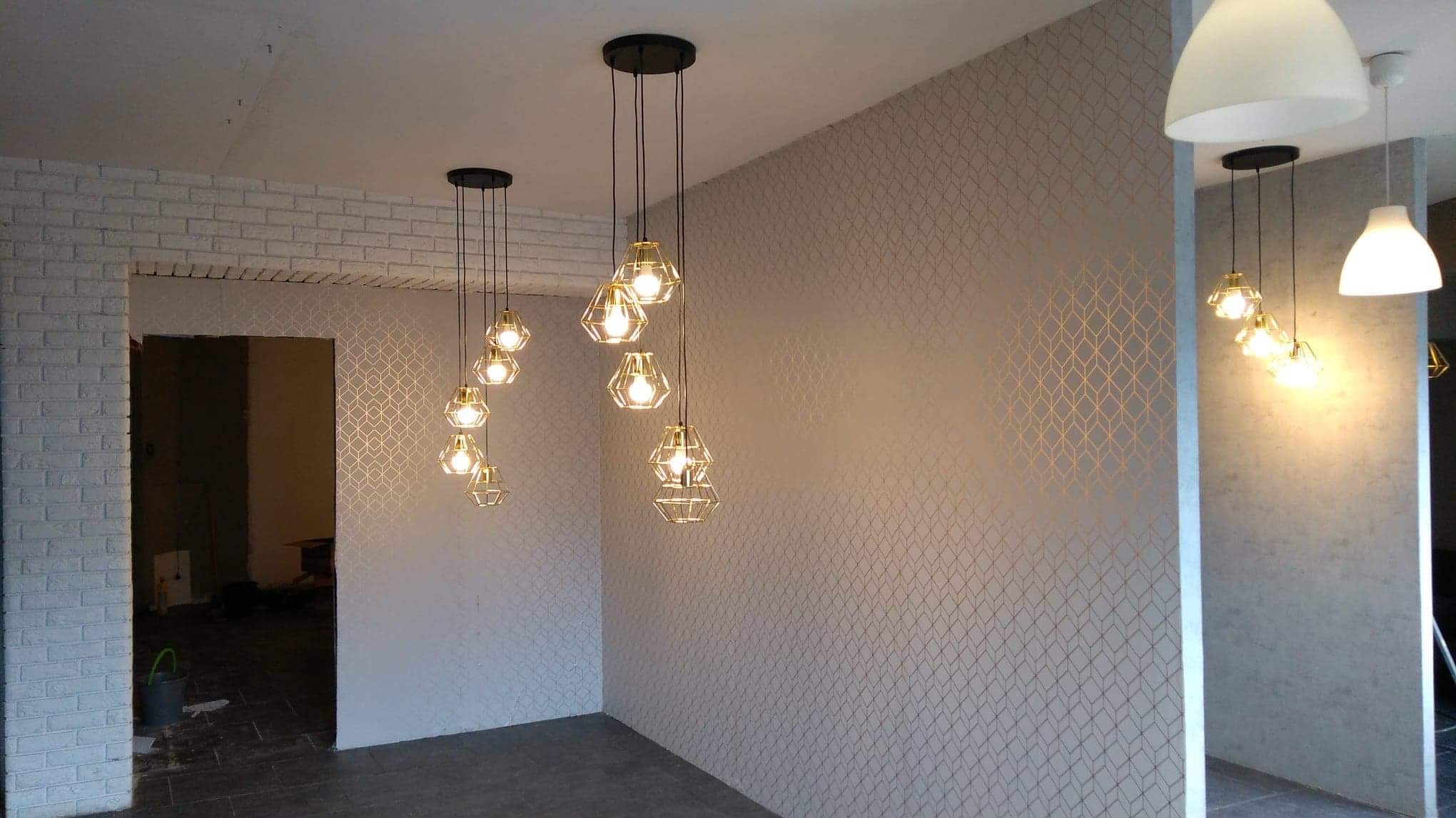 Wykończone wnętrze z dekoracyjną tapetą w geometryczny wzór i nowoczesnym oświetleniem. Widoczny fragment ściany z cegły i otwór drzwiowy.
