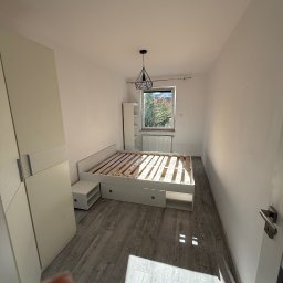 MattBud - Jasne wnętrze pokoju po odświeżeniu: białe ściany, szara podłoga, łóżko bez materaca, szafa z przesuwnymi drzwiami i okno z widokiem na zieleń. Minimalistyczny wystrój.