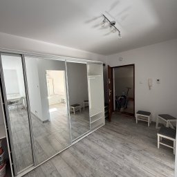 MattBud - Jasne wnętrze po odświeżeniu: duża szafa z lustrami, szara podłoga imitująca drewno, widok na inne pomieszczenia. Minimalistyczny design, nowoczesne oświetlenie.