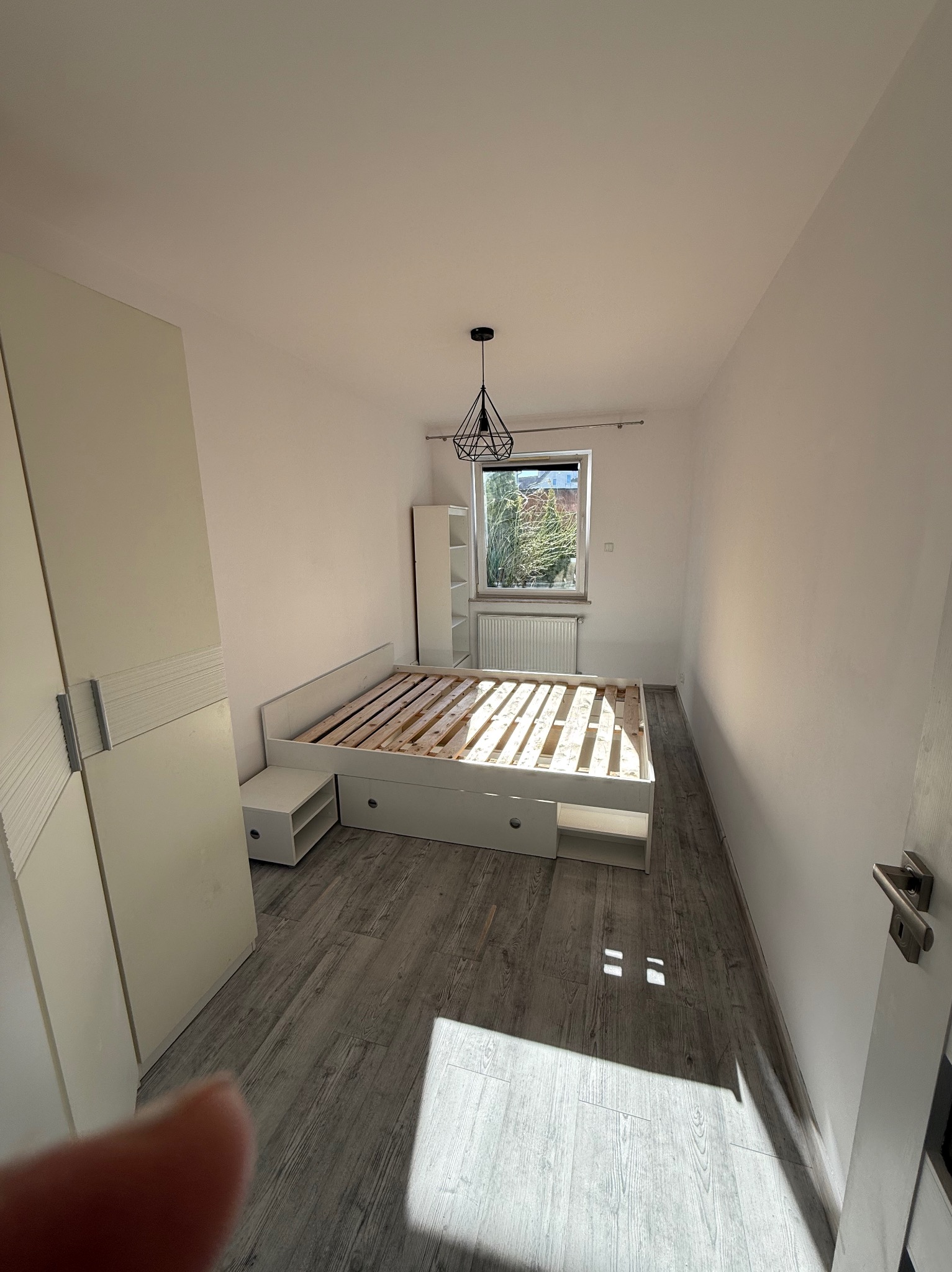 Jasne wnętrze pokoju po odświeżeniu: białe ściany, szara podłoga, łóżko bez materaca, szafa z przesuwnymi drzwiami i okno z widokiem na zieleń. Minimalistyczny wystrój.