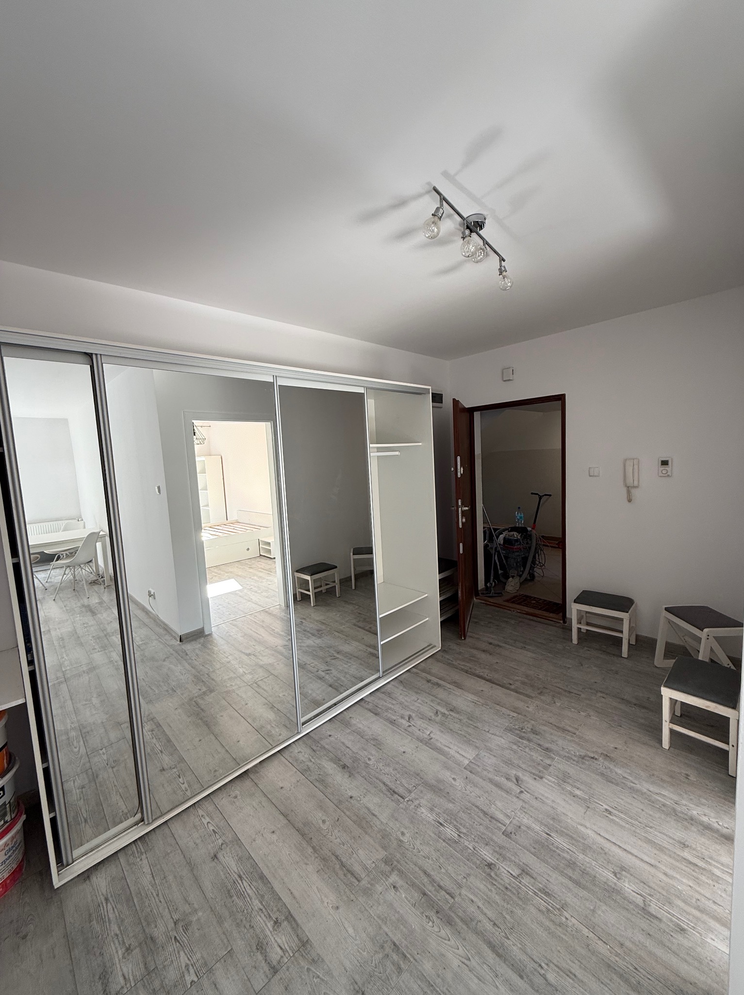 Jasne wnętrze po odświeżeniu: duża szafa z lustrami, szara podłoga imitująca drewno, widok na inne pomieszczenia. Minimalistyczny design, nowoczesne oświetlenie.