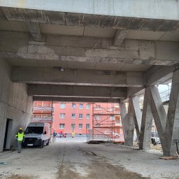 Przemysław Przychodzki - Surowa konstrukcja żelbetowa w trakcie budowy, widoczne wsporniki, szalunki i pracownicy w tle. Elementy rusztowań po prawej stronie kadru.