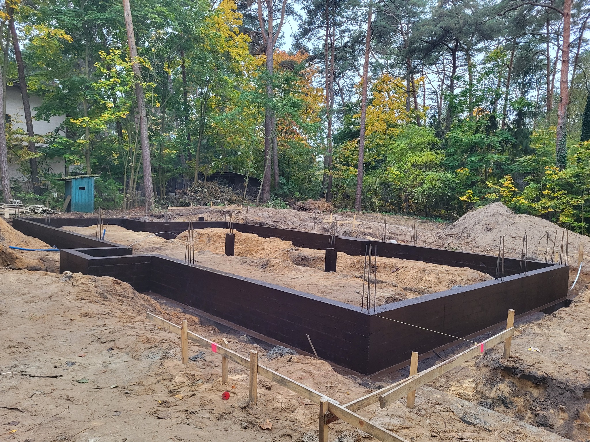 Zabezpieczone, pomalowane fundamenty domu jednorodzinnego w wykopie, z widocznym zbrojeniem i drewnianym szalunkiem. W tle drzewa i mała budka.