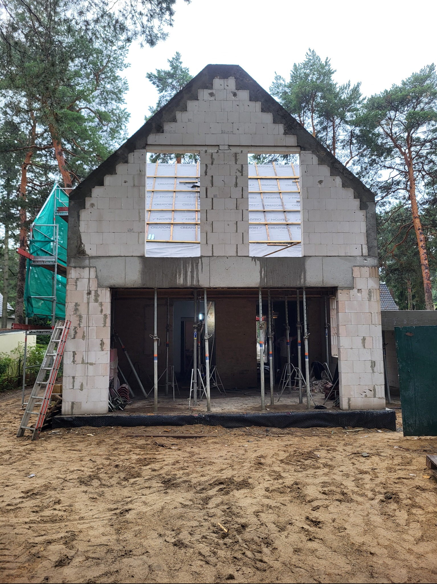 Surowy stan domu z betonu komórkowego w trakcie budowy. Widoczne podpory, rusztowania i materiały izolacyjne w otworach okiennych. Tło stanowią drzewa.