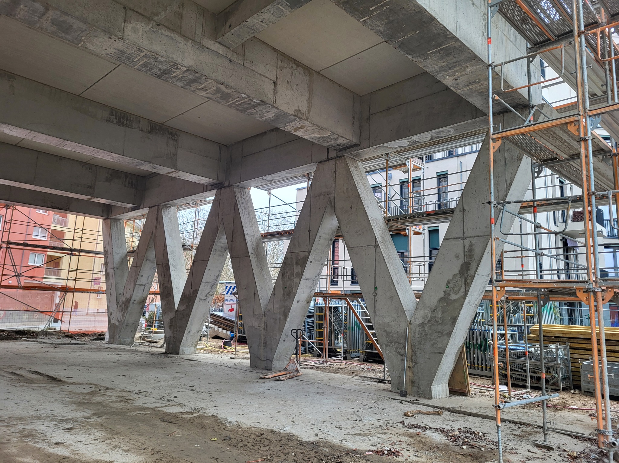 Surowa konstrukcja żelbetowa z charakterystycznymi, skośnymi podporami w trakcie budowy. Widoczne rusztowania i materiały budowlane. W tle budynki mieszkalne.