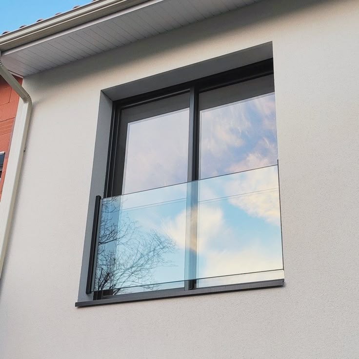 Szklana balustrada balkonowa z czarną ramą, zamontowana na elewacji budynku. W szybach odbija się błękitne niebo z chmurami i korona drzewa.