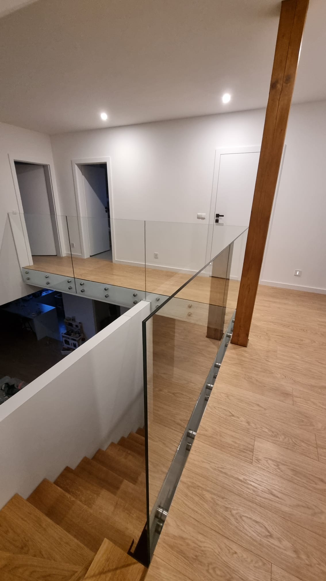 Szklana balustrada z mocowaniem punktowym w nowoczesnym wnętrzu. Widok na schody i drewnianą podłogę. Minimalistyczny design z drewnianym słupem.