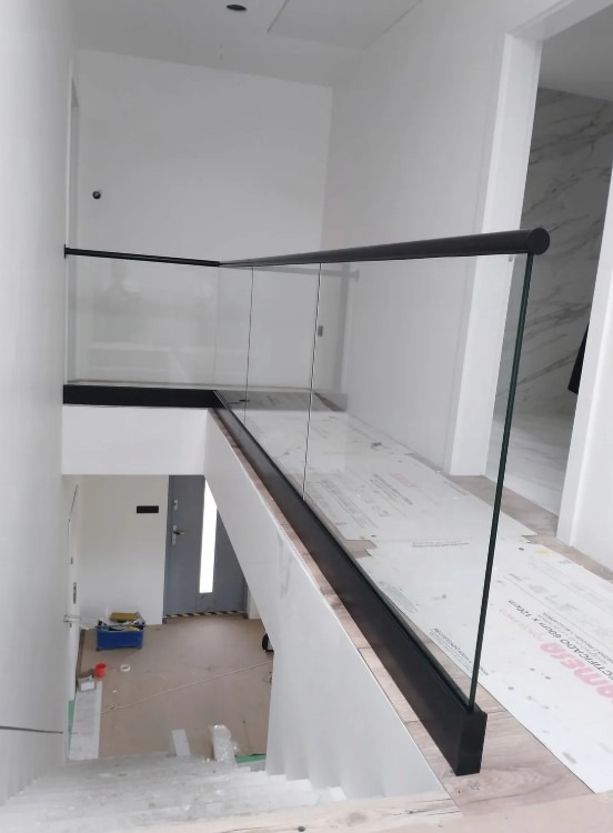Nowoczesna balustrada szklana z czarnymi profilami na antresoli. Widok z góry na schody i zabezpieczone podłoże. Minimalistyczny design, jasne wnętrze.