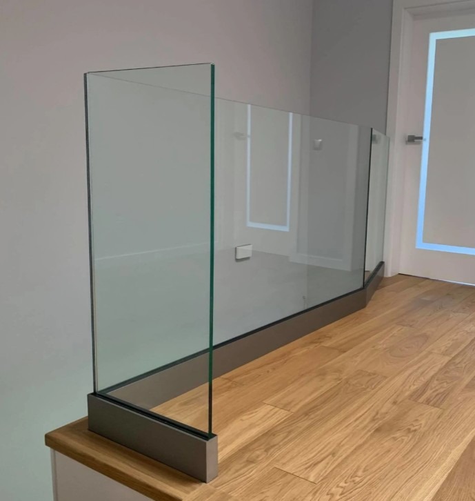 Szklana balustrada z szarą podstawą, zamontowana na drewnianych schodach w jasnym wnętrzu. Minimalistyczny design, widoczne mocowania szkła.