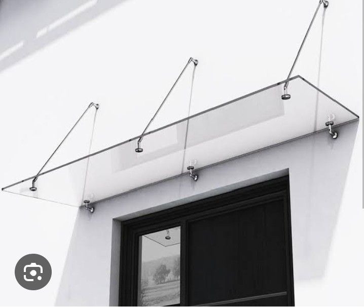 Szklany daszek nad wejściem, wsparty na metalowych linkach i mocowaniach. Minimalistyczny design, widok z dołu, czarne drzwi w tle, jasna elewacja.