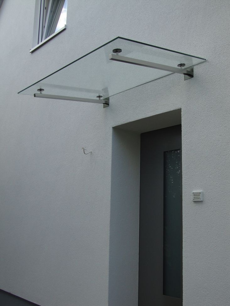 Szklany daszek nad wejściem do budynku, wsparty na metalowych wspornikach. Minimalistyczny design, szara ściana z fakturą, widoczne okno na górze.