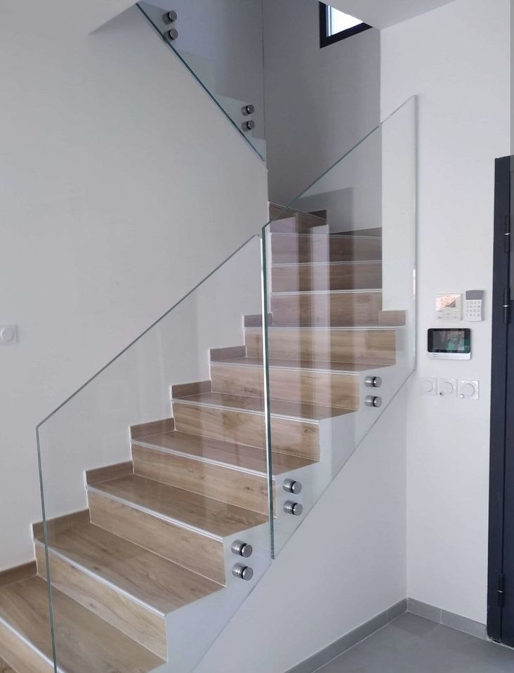 Nowoczesne schody z drewnopodobnymi stopniami i szklaną balustradą mocowaną punktowo. Minimalistyczny design, jasne wnętrze, widok z dołu na górę.