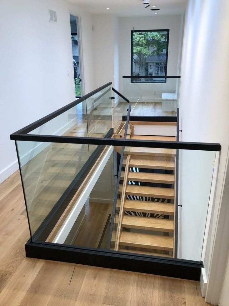 Nowoczesna balustrada szklana z czarnymi profilami, zabezpieczająca schody w jasnym wnętrzu. Widok z góry na drewniane schody i podłogę.