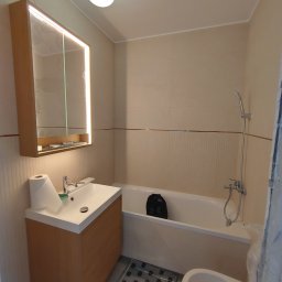 YEVHEN SVIRIDOV - Jasna łazienka po remoncie: beżowe płytki, podświetlane lustro nad umywalką z szafką, wanna z prysznicem, toaleta. Na podłodze mata ochronna.