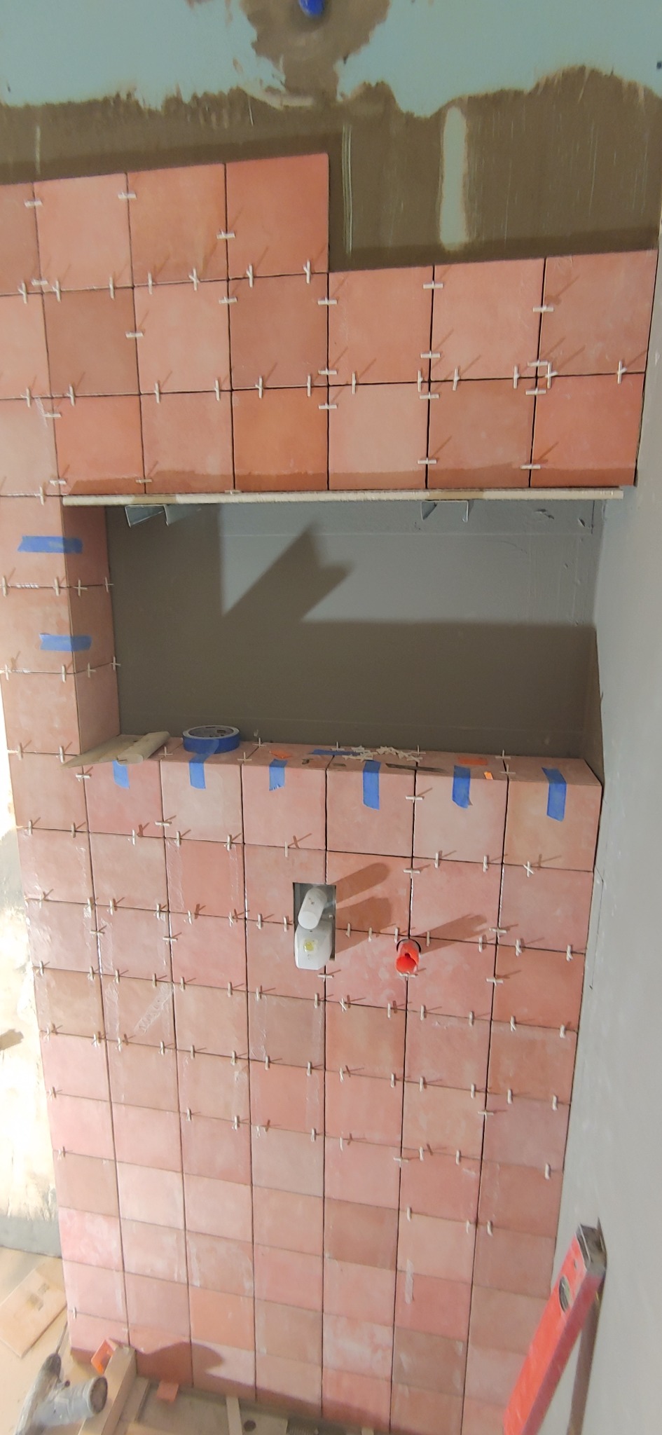 Ściana z terakotowych płytek w trakcie układania, z widocznymi krzyżykami dystansowymi i niebieską taśmą maskującą, tworząca wnękę. Widoczne narzędzia glazurnicze.
