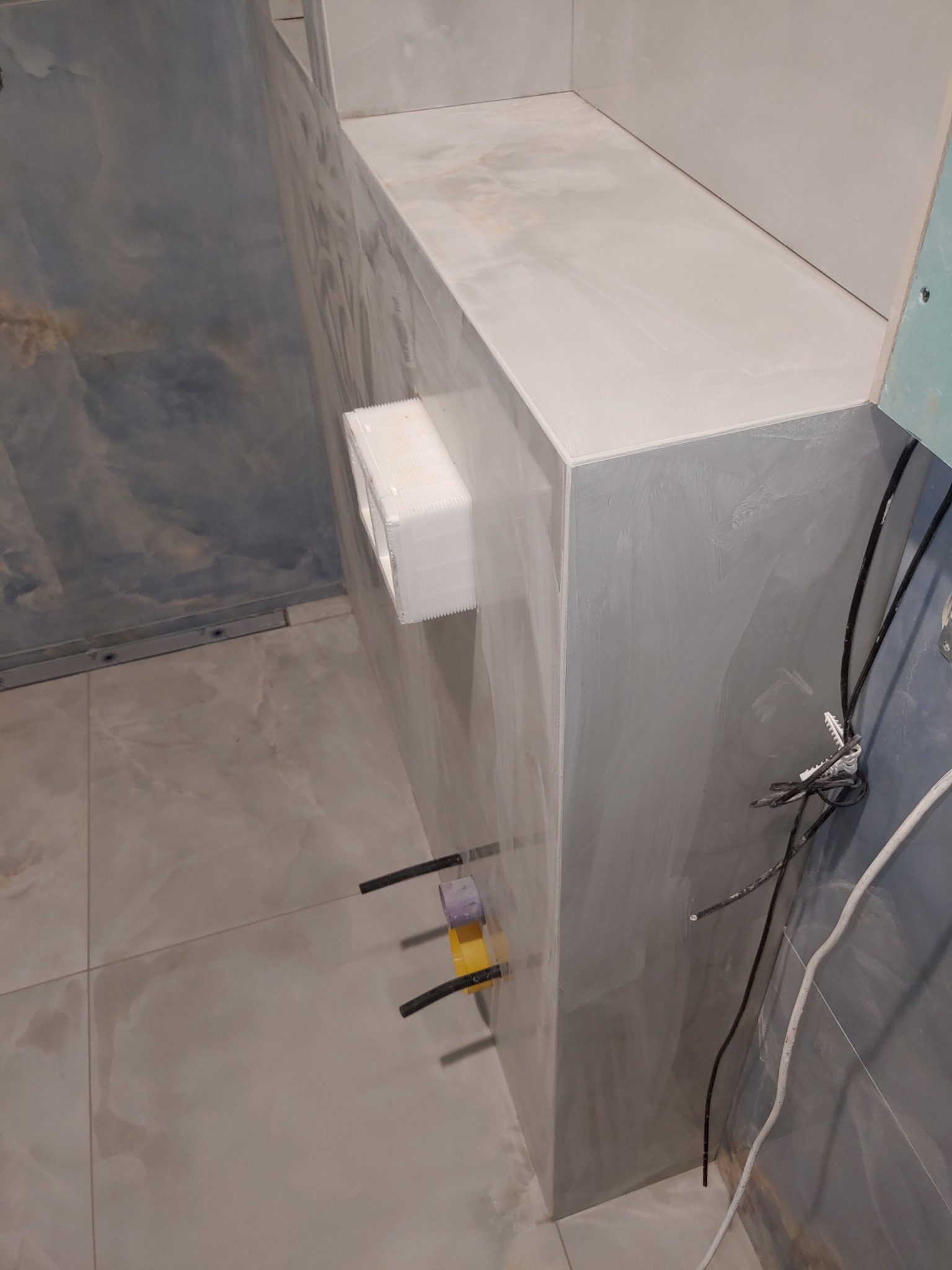 Szara, marmurkowa glazura na ścianie i podłodze łazienki. Widoczne elementy instalacji hydraulicznej i elektrycznej przed ostatecznym wykończeniem.