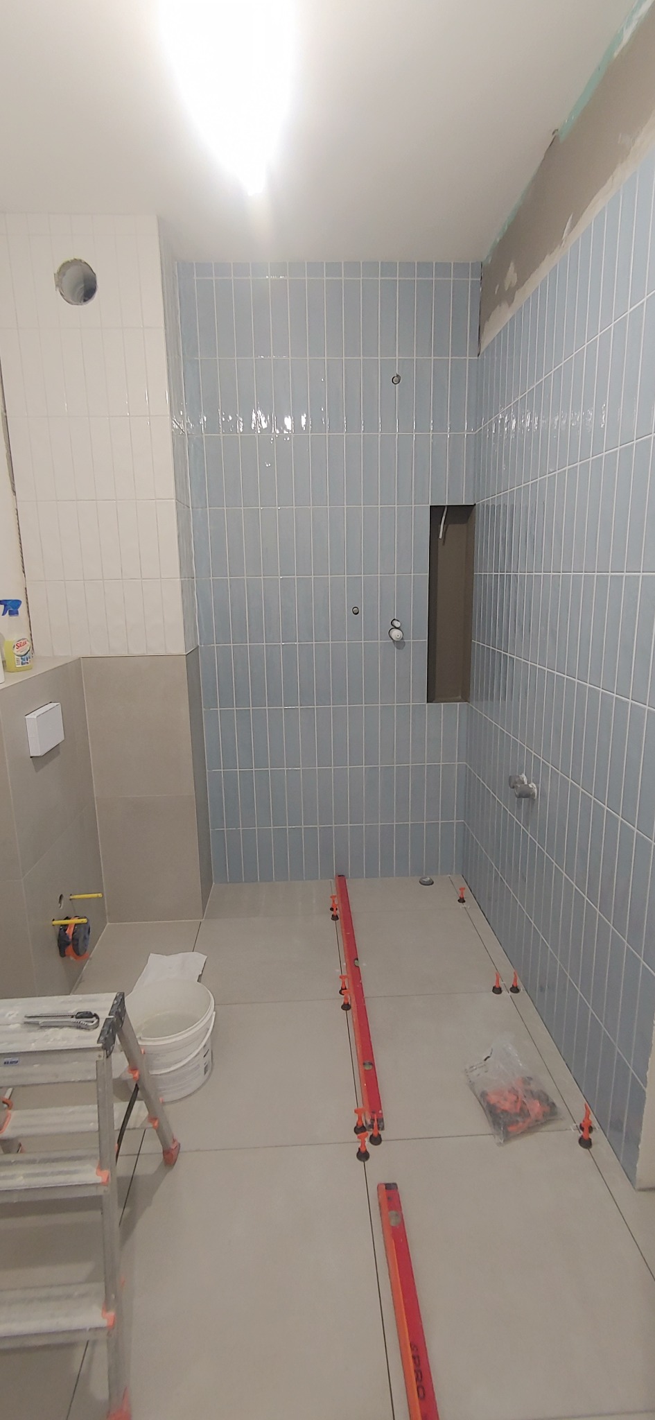 Łazienka w trakcie remontu: błękitne płytki na ścianach, poziomice i system poziomowania na podłodze. Widoczny fragment drabiny i wiadro. Oświetlenie sufitowe.