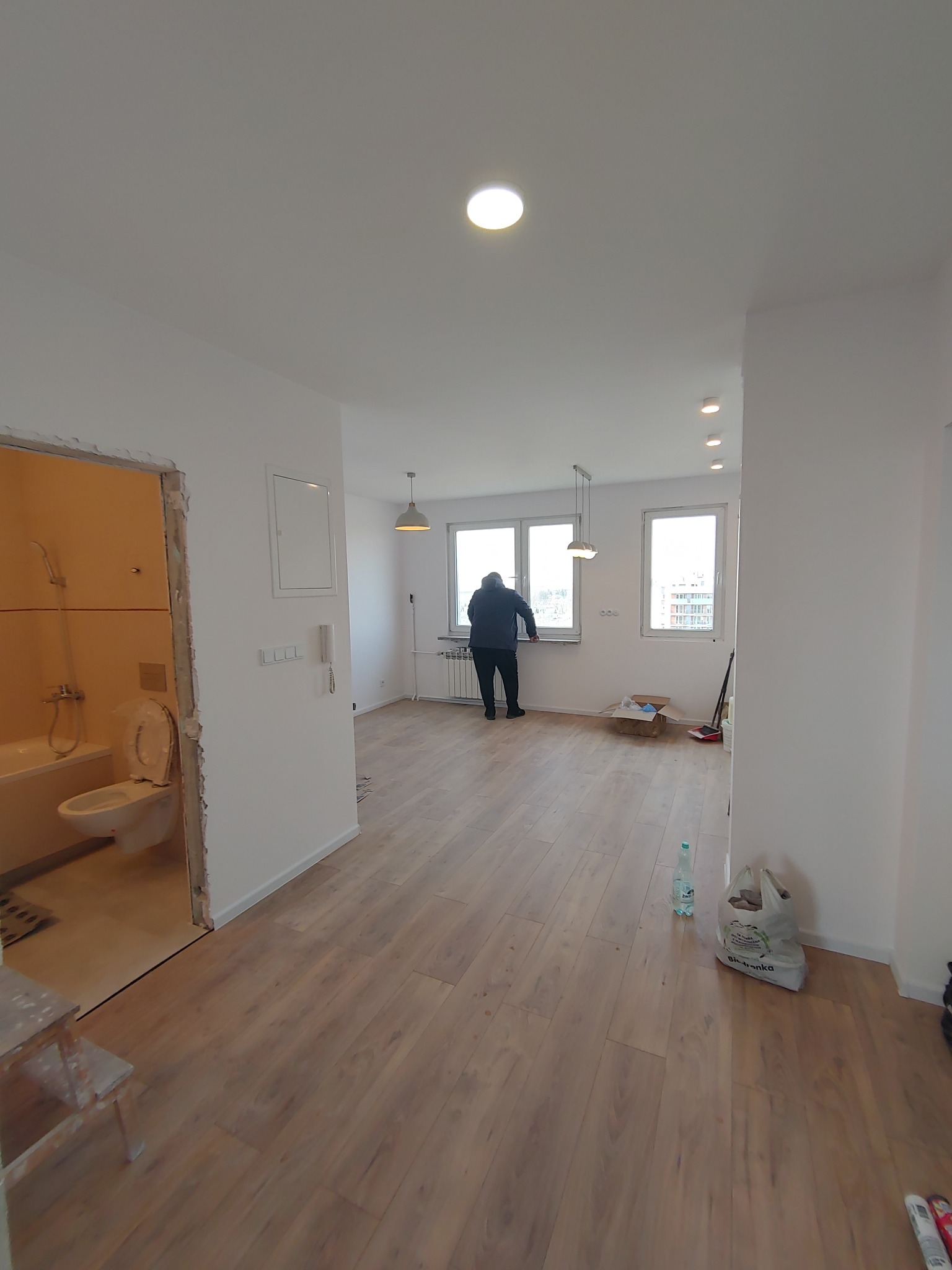 Jasne panele podłogowe w wyremontowanym mieszkaniu w Warszawie. Widoczna łazienka w trakcie prac i osoba przy oknie. Minimalistyczne lampy sufitowe.