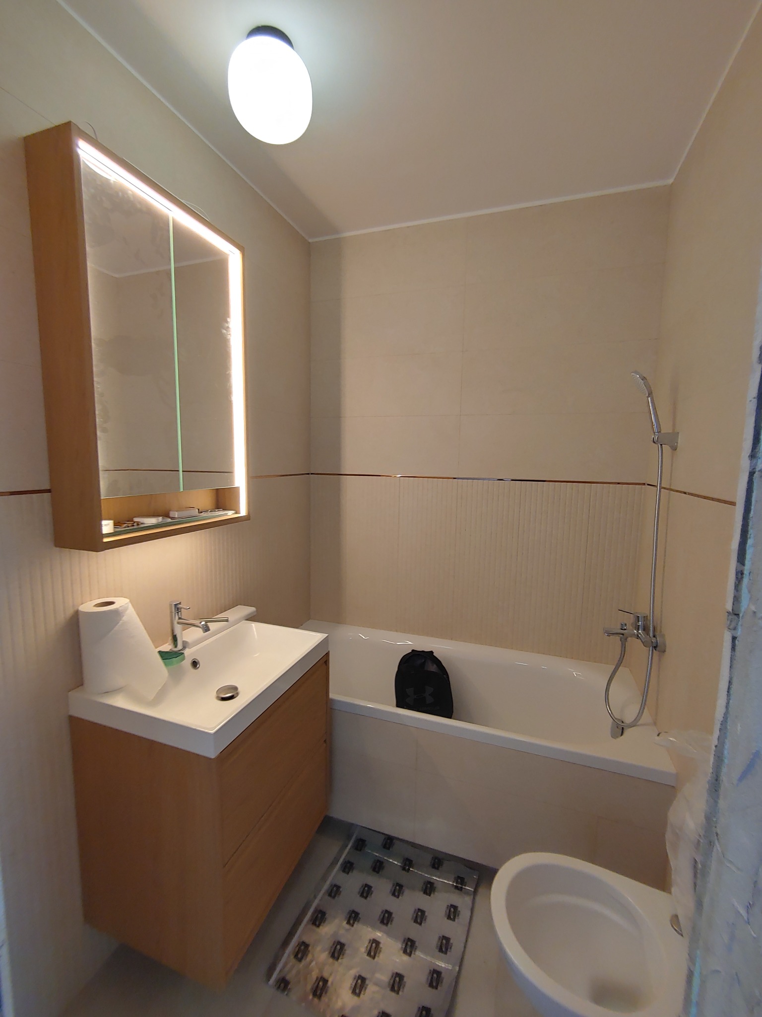 Jasna łazienka po remoncie: beżowe płytki, podświetlane lustro nad umywalką z szafką, wanna z prysznicem, toaleta. Na podłodze mata ochronna.