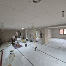 Hubert Siepak - Wnętrze w trakcie prac wykończeniowych: ściany i sufit z płyt kartonowo-gipsowych, widoczny pracownik przygotowujący materiały, narzędzia i drabina w tle.