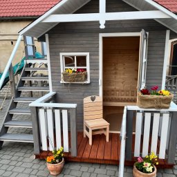 Hubert Siepak - Drewniany domek dla dzieci z schodami, zjeżdżalnią i małym tarasem. Szare ściany, czerwony dach, kwiaty w doniczkach. Krzesło z sercem na werandzie.