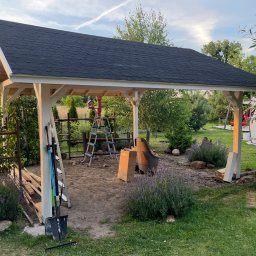 Hubert Siepak - Zadaszona konstrukcja z czarnym dachem z gontu bitumicznego i białymi słupami, w trakcie budowy, z narzędziami i materiałami budowlanymi w tle ogrodu.