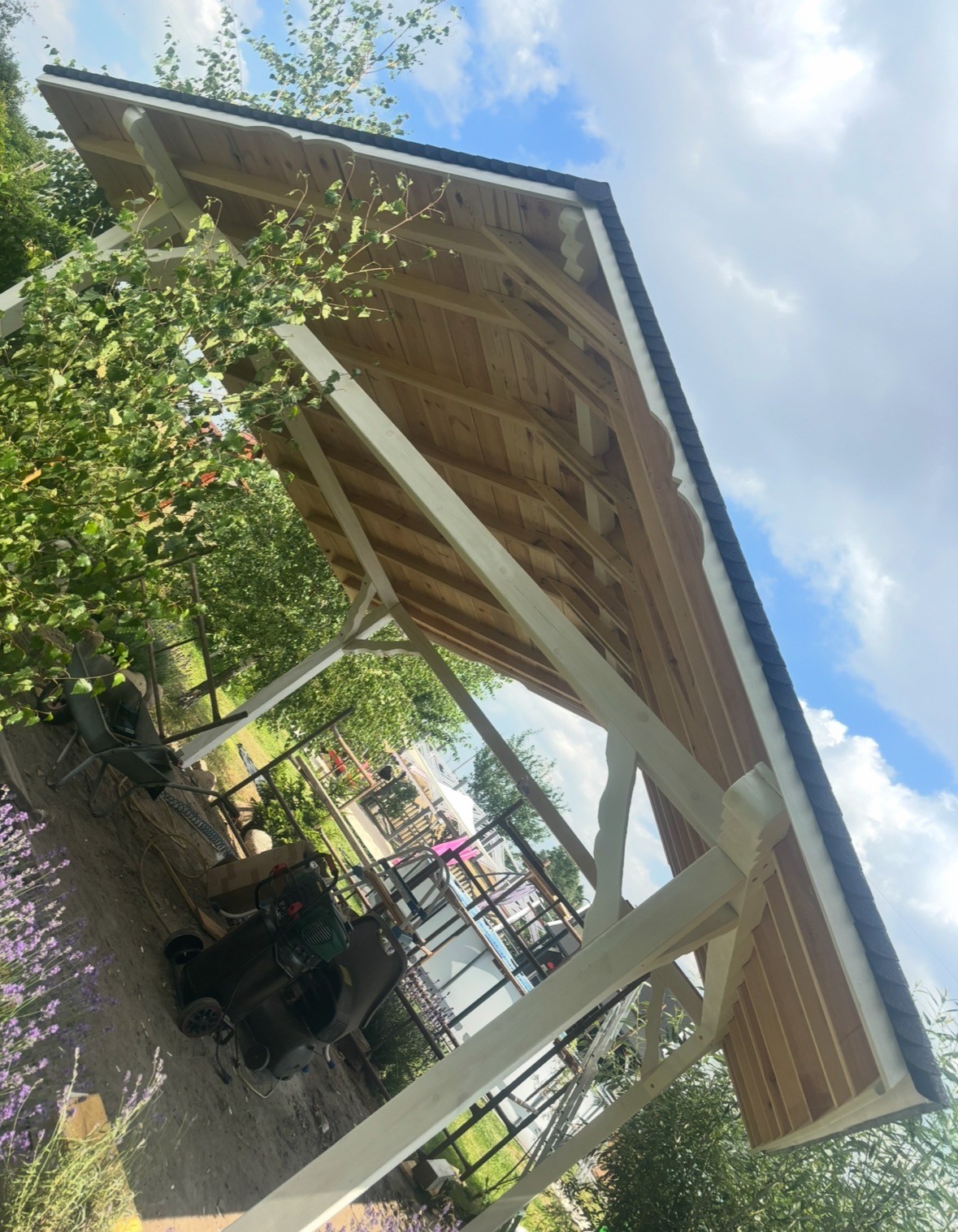 Drewniana konstrukcja dachu z widoczną więźbą, białe elementy nośne, czarne wykończenie dachu, zielone otoczenie, niebo z chmurami w tle. Ujęcie z dołu, pod kątem.