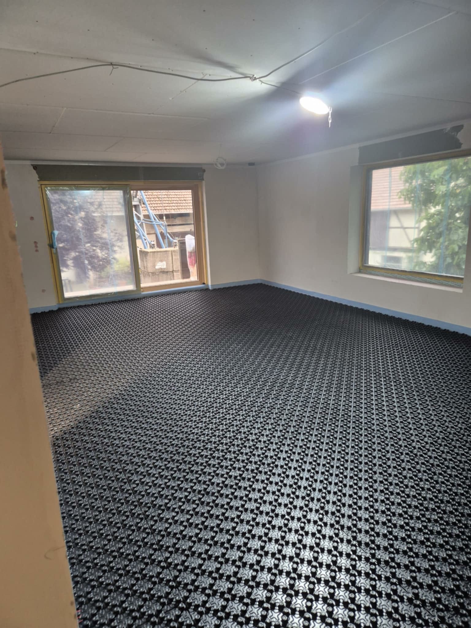 Czarna mata grzewcza pod podłogówkę, rozłożona w jasnym pokoju z oknami. Widoczne przygotowania do wylewki. Taśma brzegowa zabezpiecza ściany.