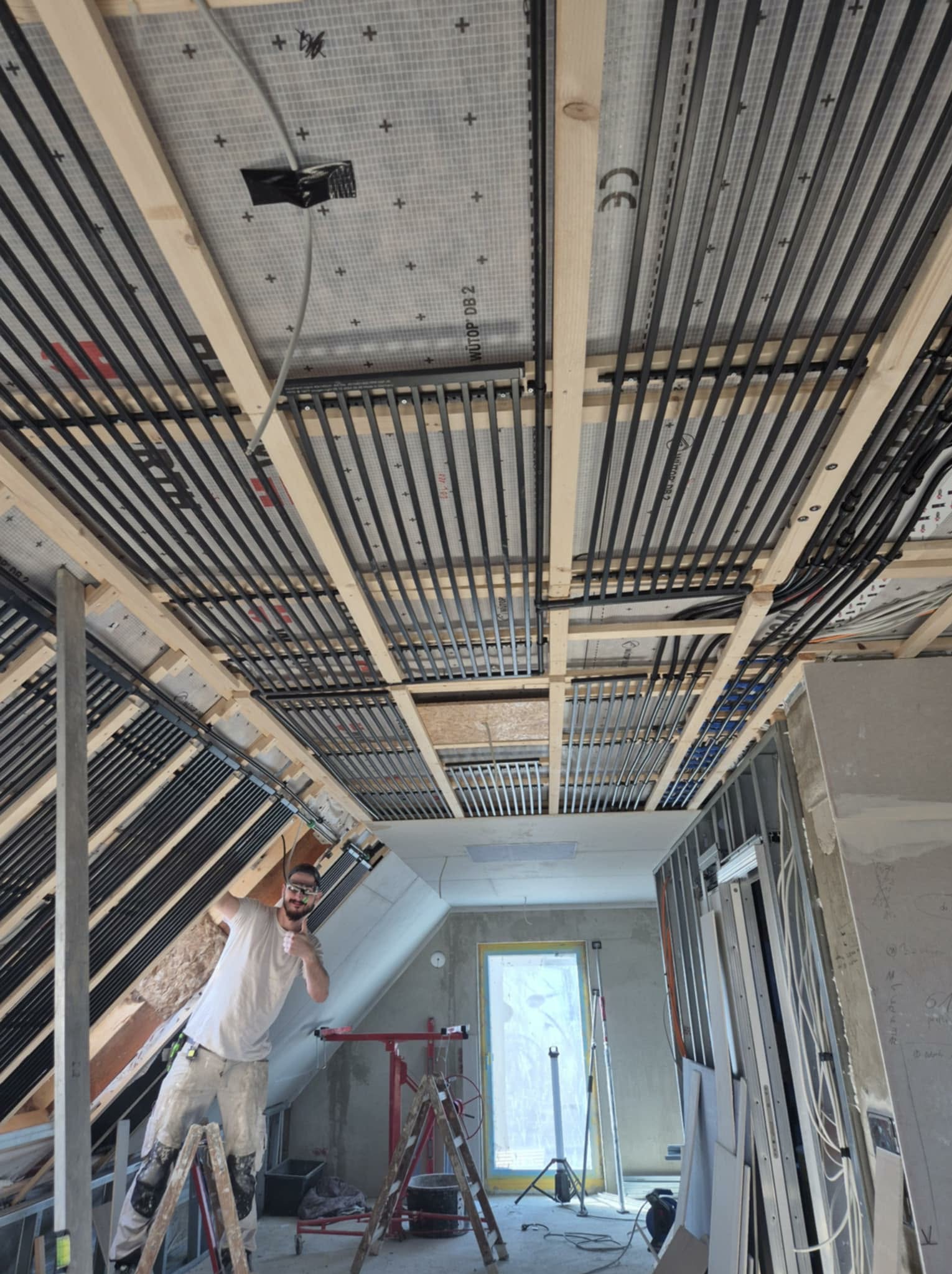 Montaż ogrzewania podłogowego na poddaszu: czarne rury grzewcze ułożone między drewnianymi belkami stropu, widoczny pracownik na drabinie, surowe wnętrze w trakcie prac.