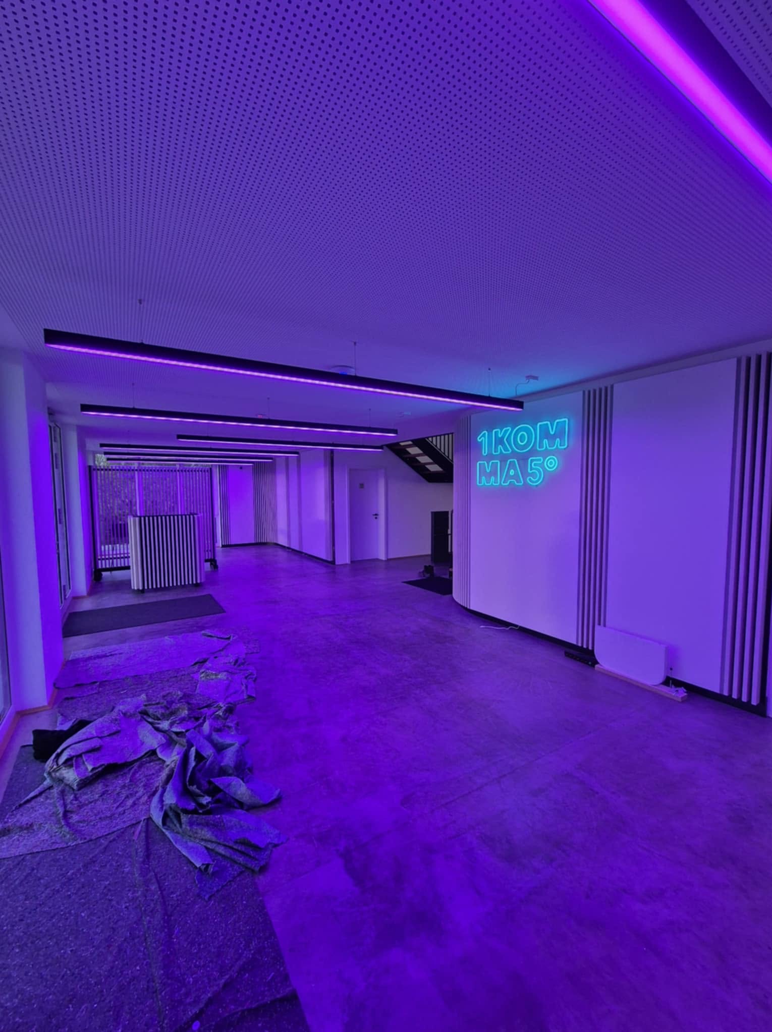Wnętrze biurowe z podwieszanym sufitem i neonem '1KOM MA 5°', oświetlone fioletowym światłem. Na podłodze rozłożone zabezpieczenia przed pracami wykończeniowymi.