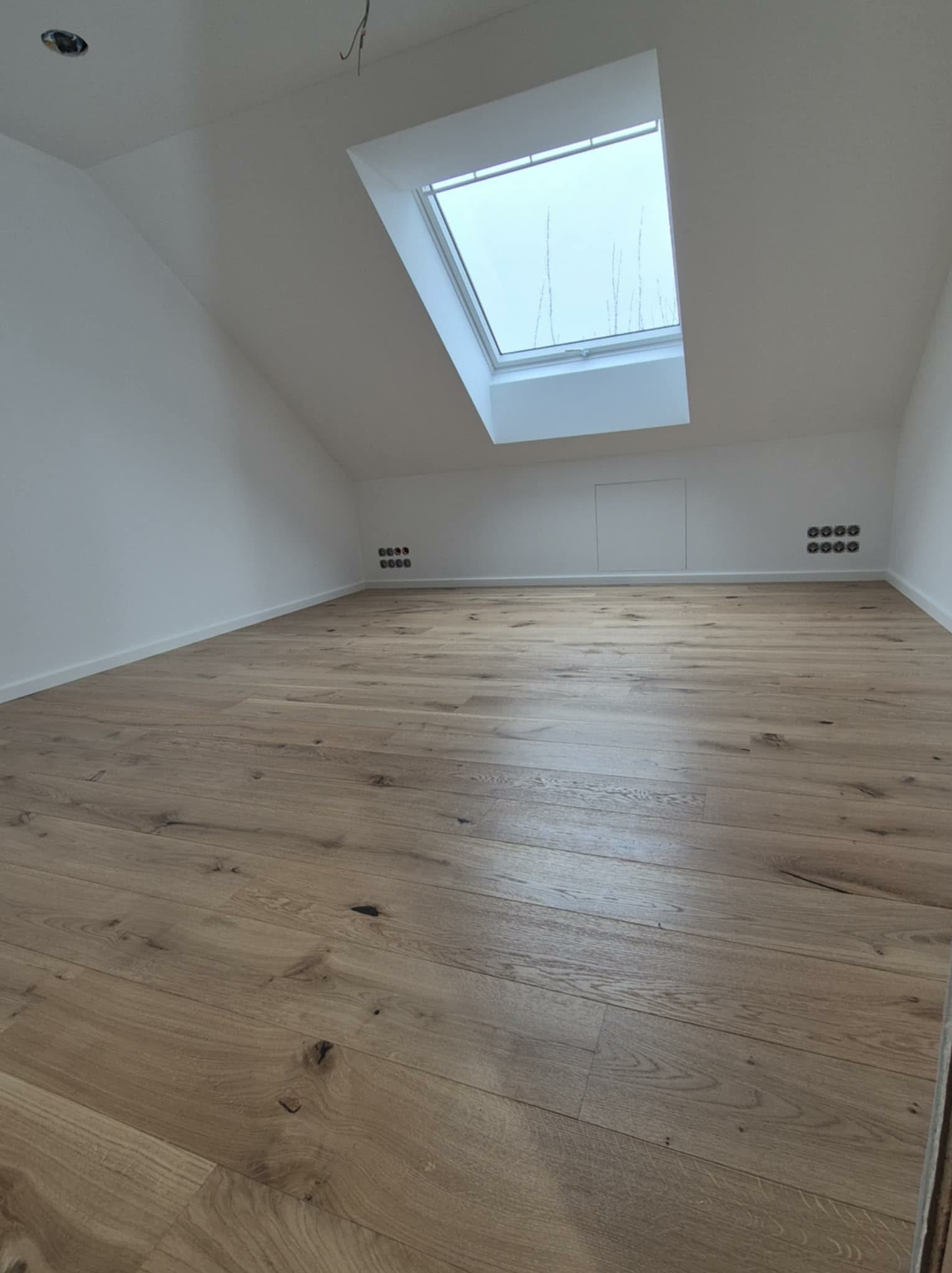 Jasne panele podłogowe w pokoju na poddaszu z oknem dachowym i białymi ścianami. Widoczne gniazdka elektryczne. Ukończona realizacja układania paneli.