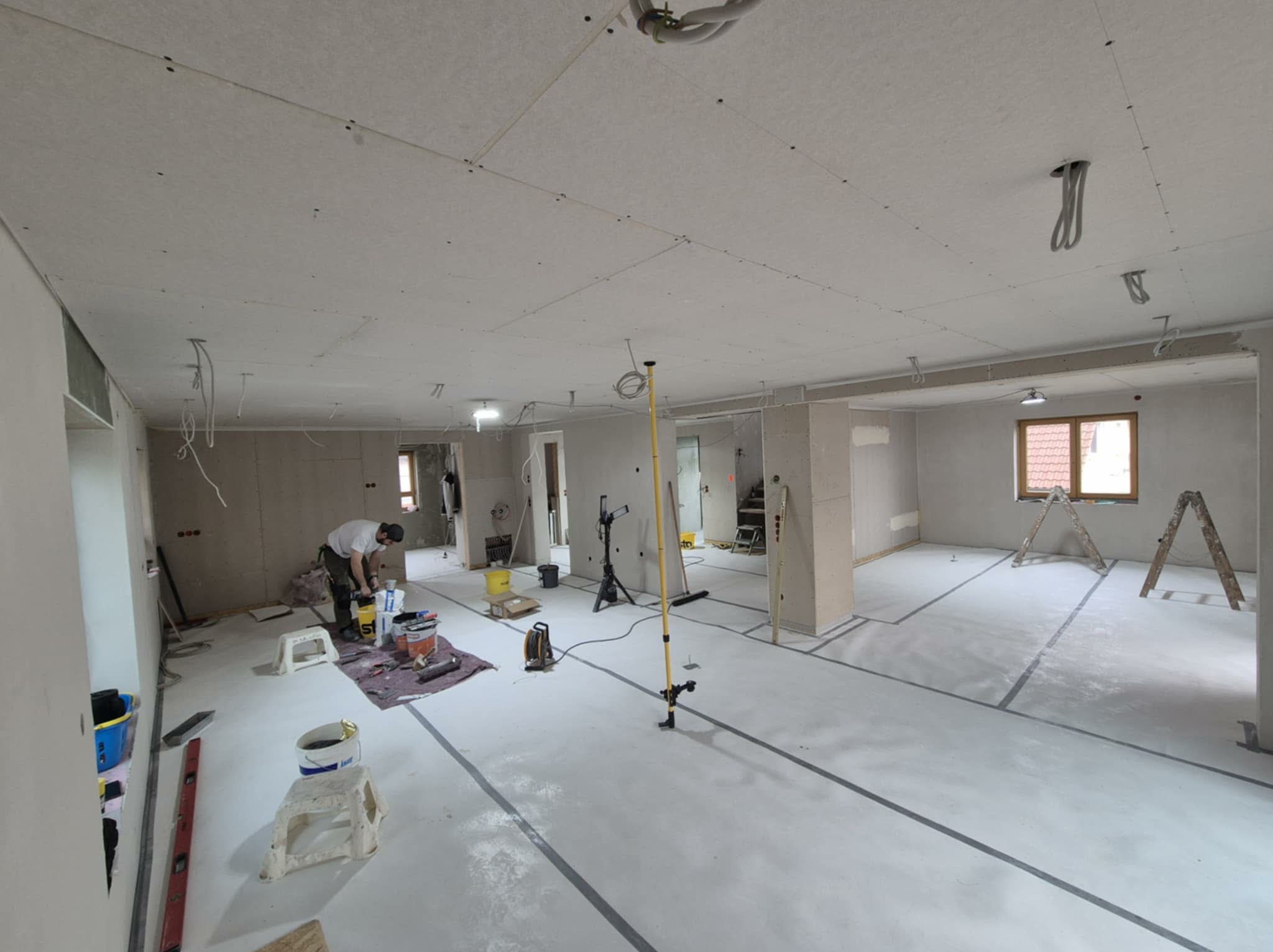 Wnętrze w trakcie prac wykończeniowych: ściany i sufit z płyt kartonowo-gipsowych, widoczny pracownik przygotowujący materiały, narzędzia i drabina w tle.