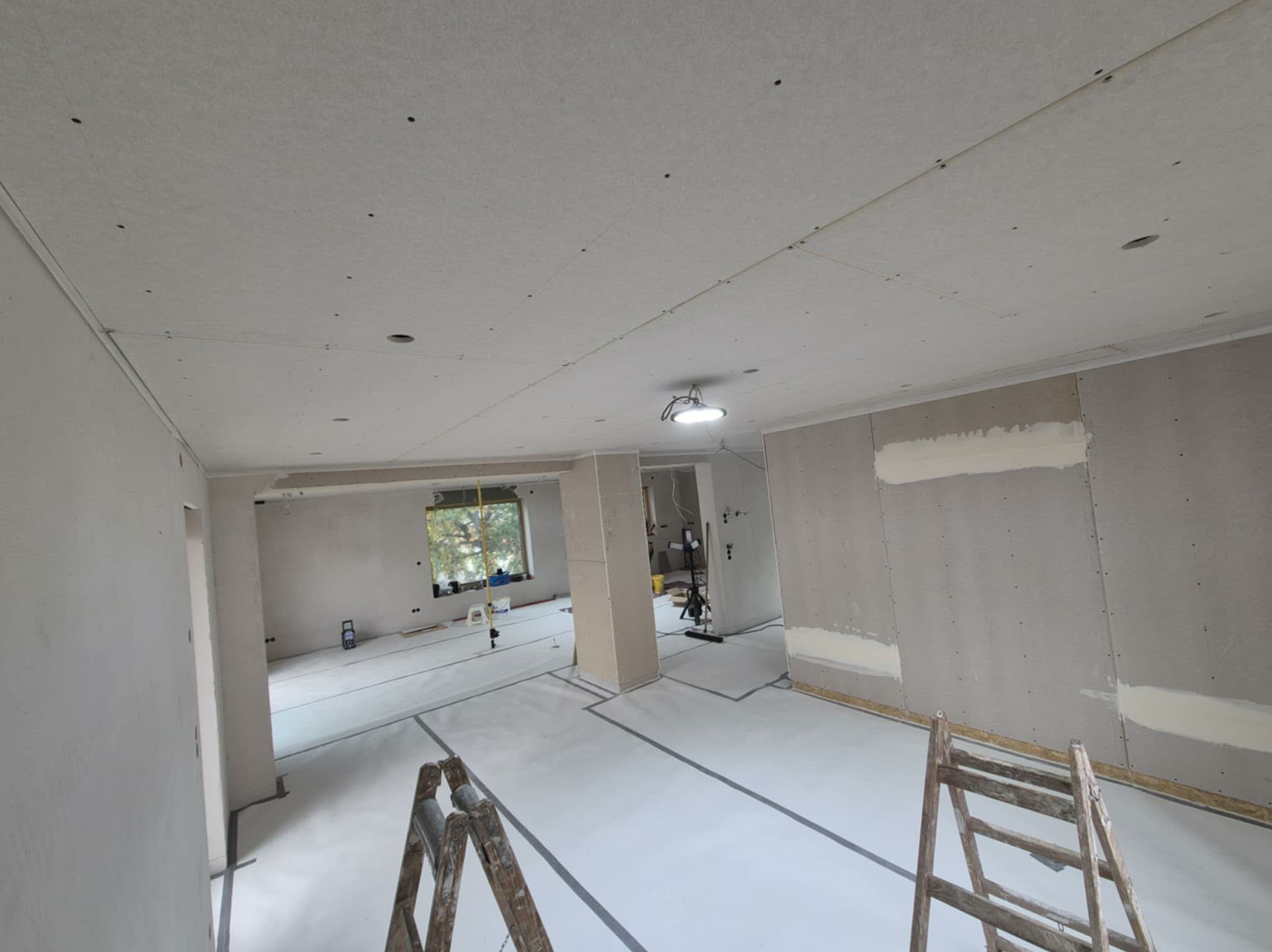 Wnętrze w trakcie prac wykończeniowych: ściany i sufit pokryte płytami kartonowo-gipsowymi, widoczne drabiny i przygotowane podłoże. Oświetlenie sufitowe.