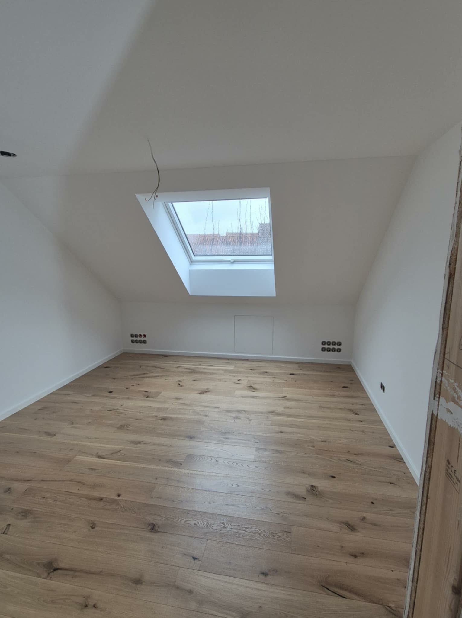 Jasne panele podłogowe w pokoju na poddaszu z oknem dachowym. Świeżo pomalowane ściany na biało. Widoczne gniazdka elektryczne. Ujęcie z poziomu podłogi.