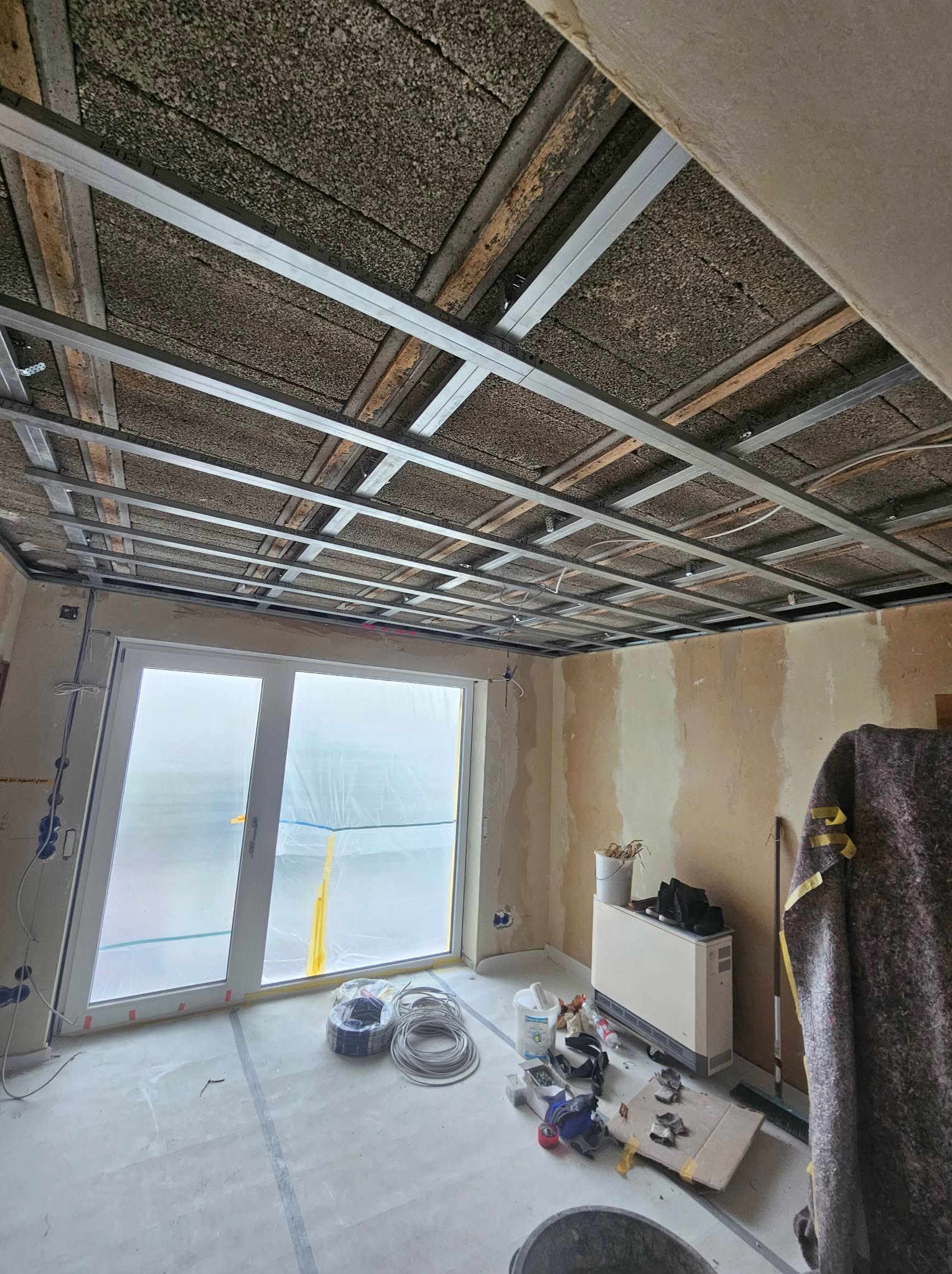 Sufit podwieszany w trakcie montażu: metalowa konstrukcja kratowa, izolacja akustyczna, okno zabezpieczone folią. Widoczne przewody i narzędzia na podłodze.