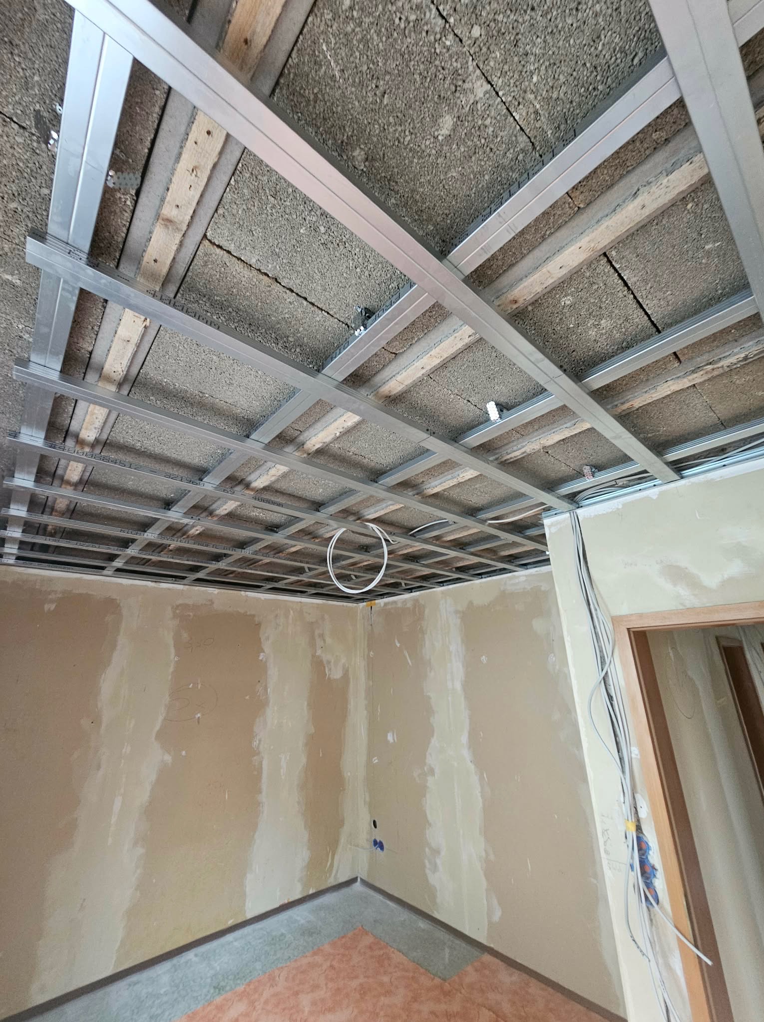 Sufit podwieszany z metalową konstrukcją w trakcie montażu. Widoczne płyty stropowe i instalacja elektryczna. Ściany przygotowane do dalszych prac wykończeniowych.