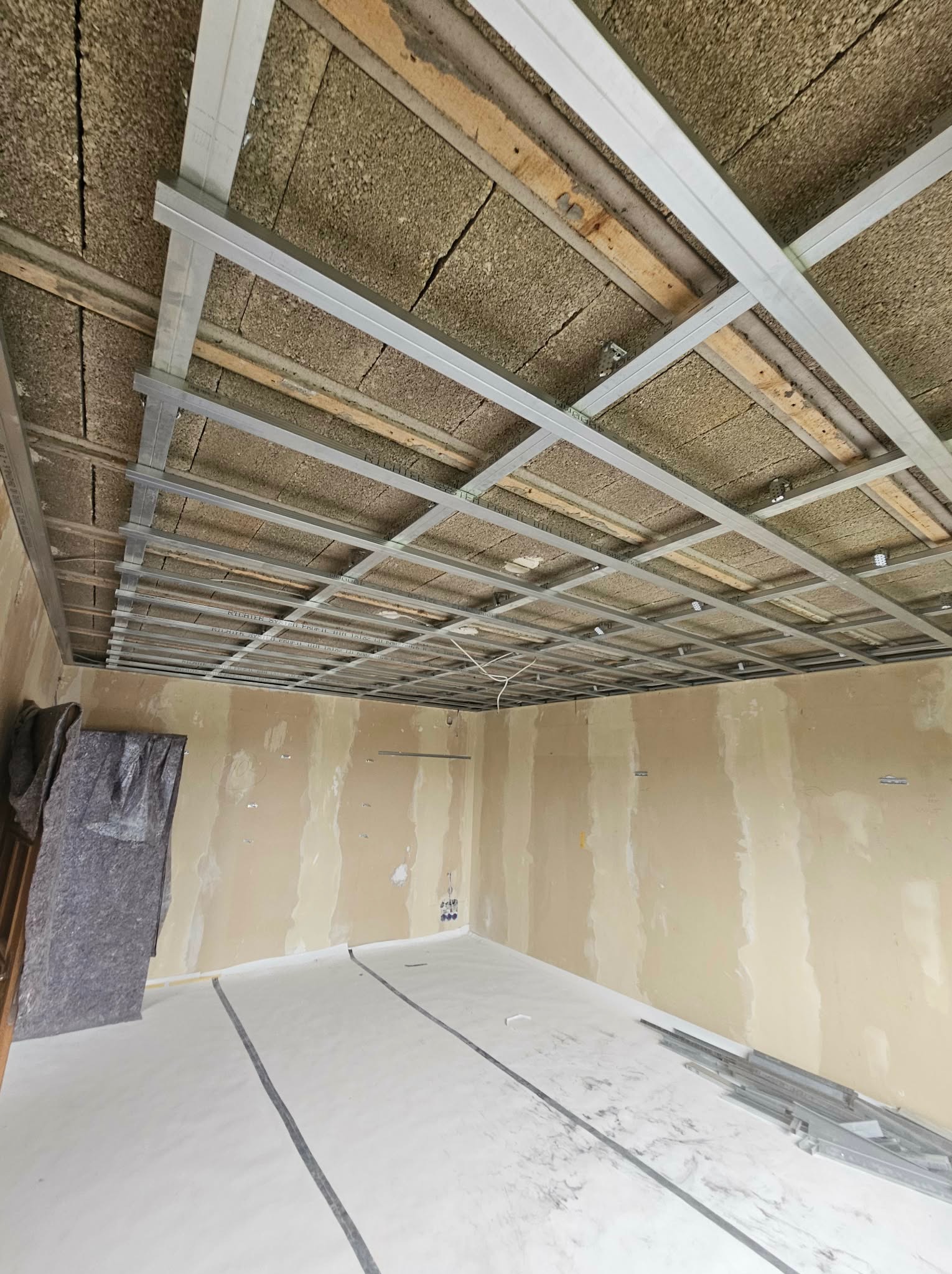 Montaż metalowej konstrukcji sufitu podwieszanego z izolacją z wełny mineralnej. Widoczne profile i płyty izolacyjne. Przygotowanie do montażu płyt karton-gips.