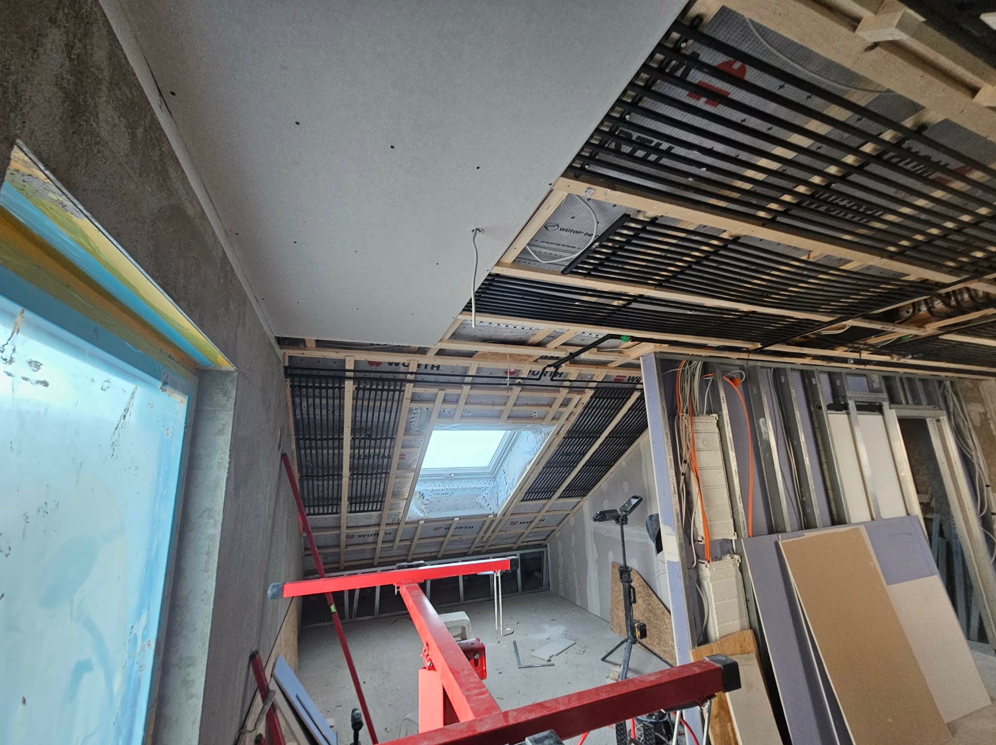 Montaż płyty KG na suficie poddasza z oknem dachowym. Widoczne profile, instalacja grzewcza sufitowa i częściowo wykończone ściany. Czerwony podnośnik do płyt.